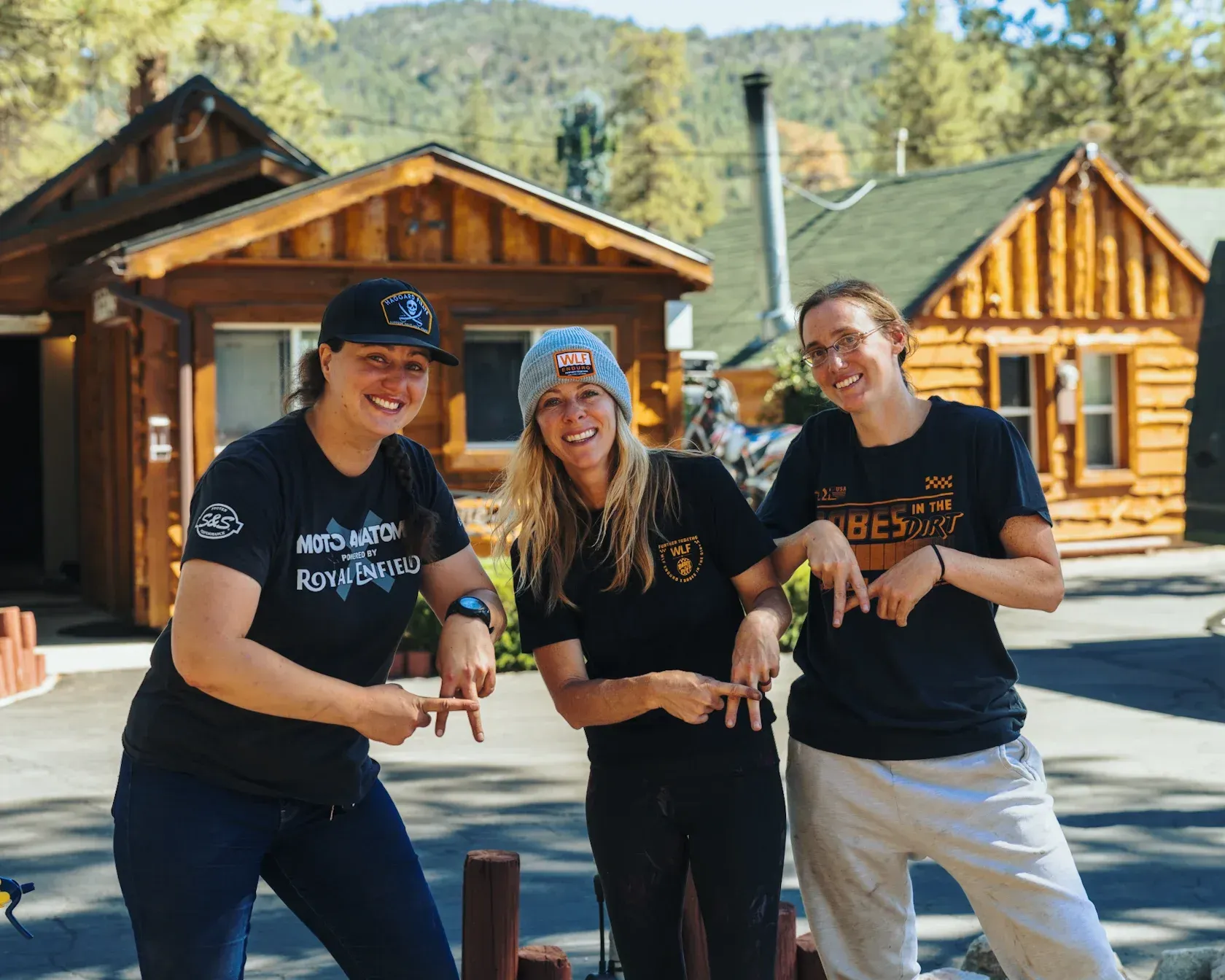 Three women smiling, pointing, in front of cabins. Sunny setting, all wearing t-shirts, one with a beanie.