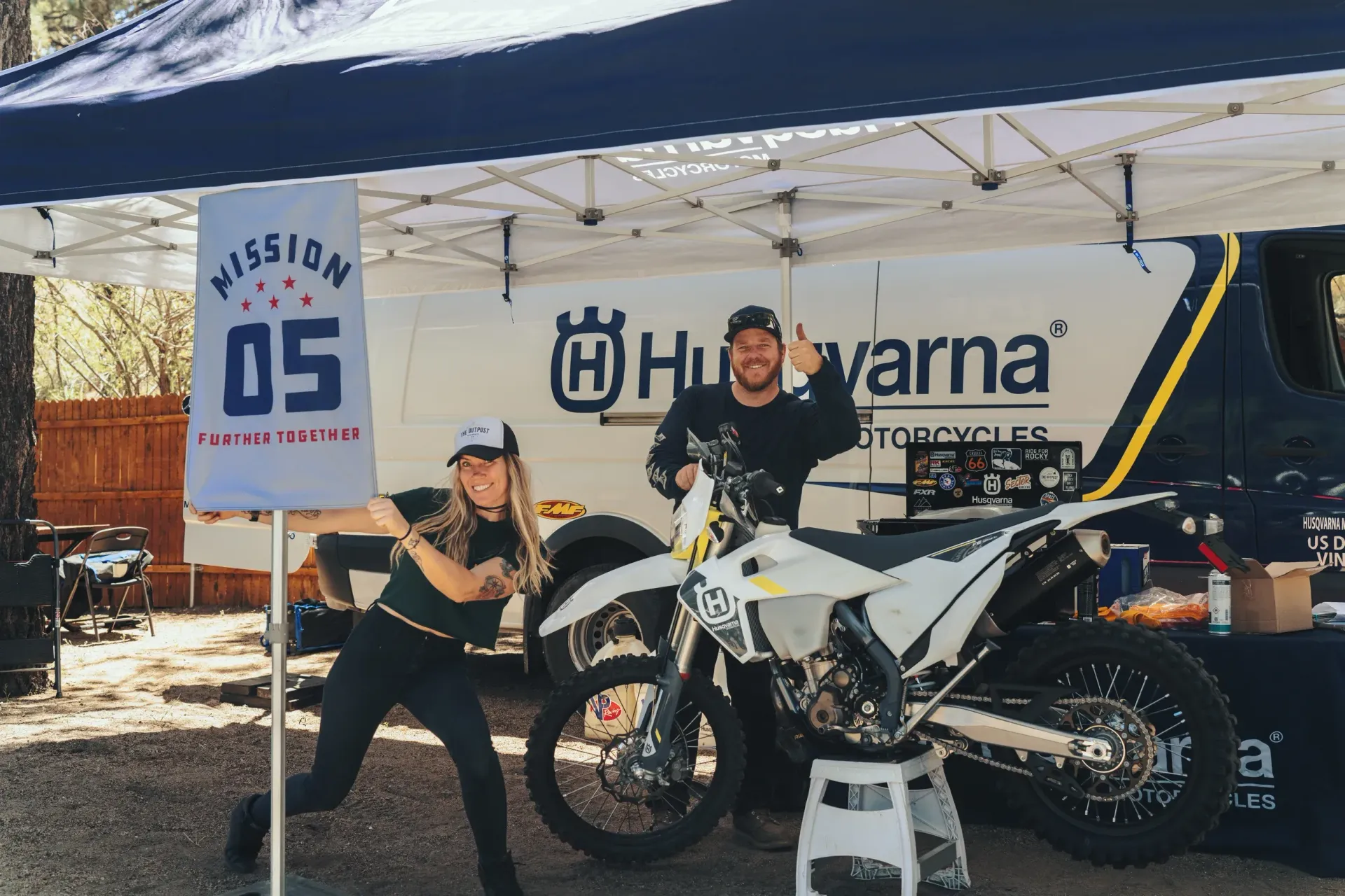 Two people with a Husqvarna motorcycle in front of a tent. Woman holds a sign; man gives a thumbs-up.