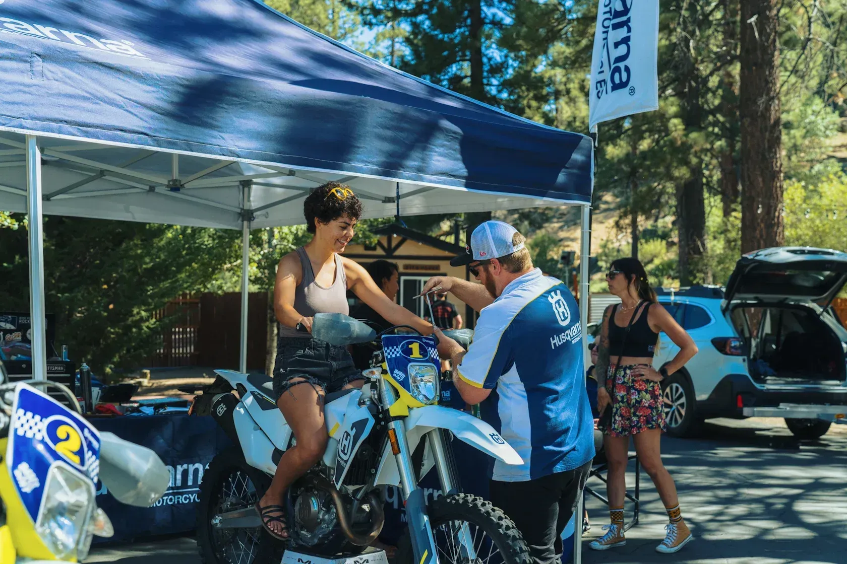 Woman on motorcycle, technician adjusting it; outdoor event with tent, flag, and parked car.