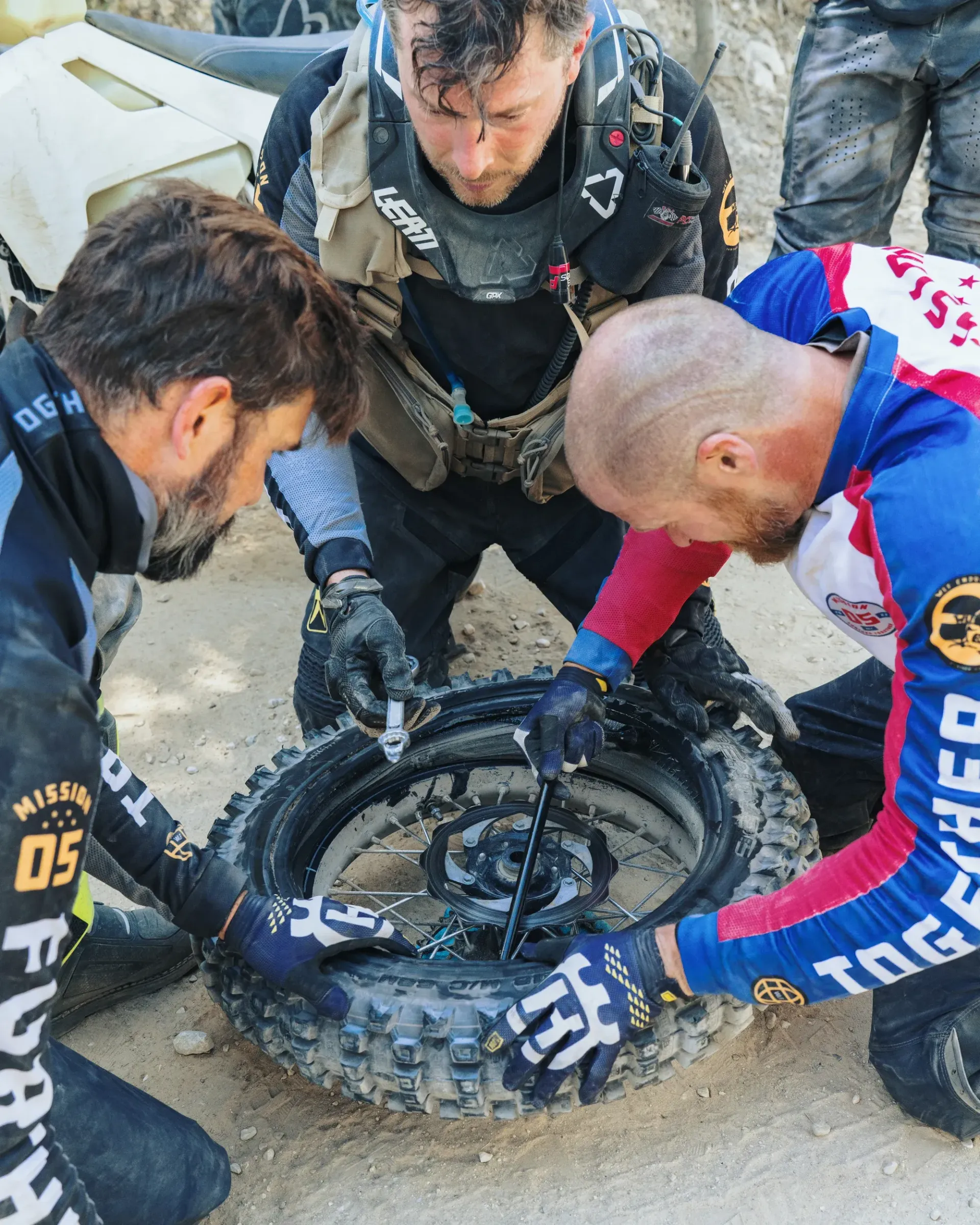 Three people fixing a motorcycle tire outdoors. They wear gloves and protective gear.