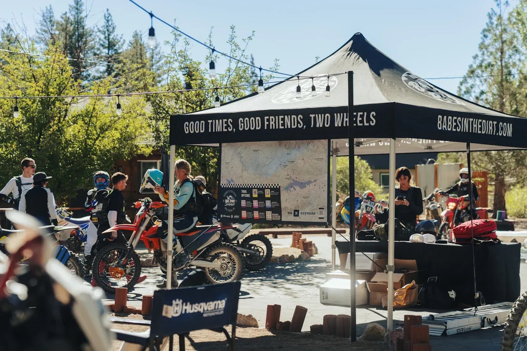 Outdoor event with motorcycles and a tent. People gather around, some looking at merchandise. Sunny day.