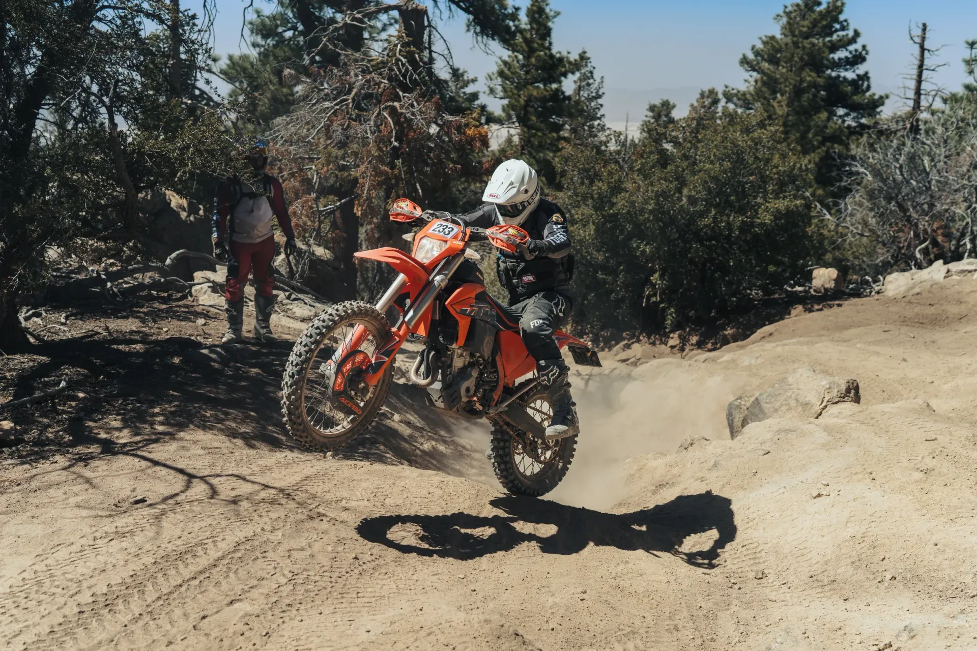 Dirt bike rider navigating a dusty trail in a forest; rider is wearing helmet and gear.