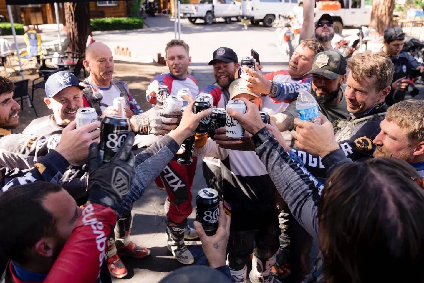Group of men in motorcycle gear cheer, holding up cans and bottles. Outdoors, sunny day.