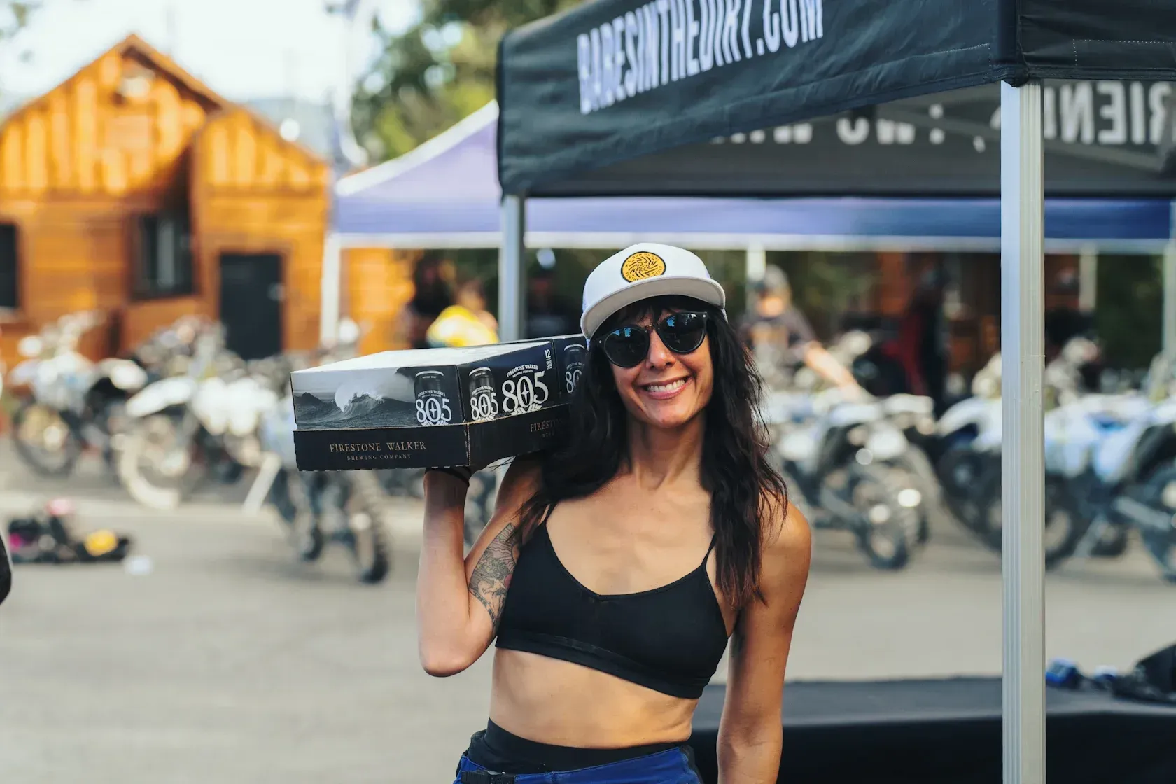 Woman in sunglasses and cap holding box, smiling, near motorcycles. Outdoors.
