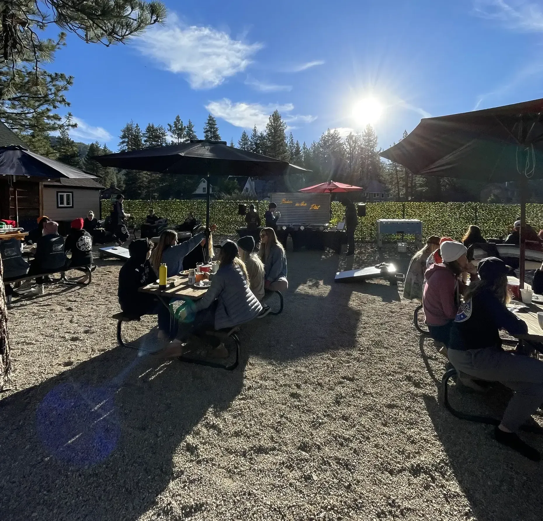 People seated at picnic tables outdoors, sunny day. Some under umbrellas, eating/drinking. Buildings, trees in background.