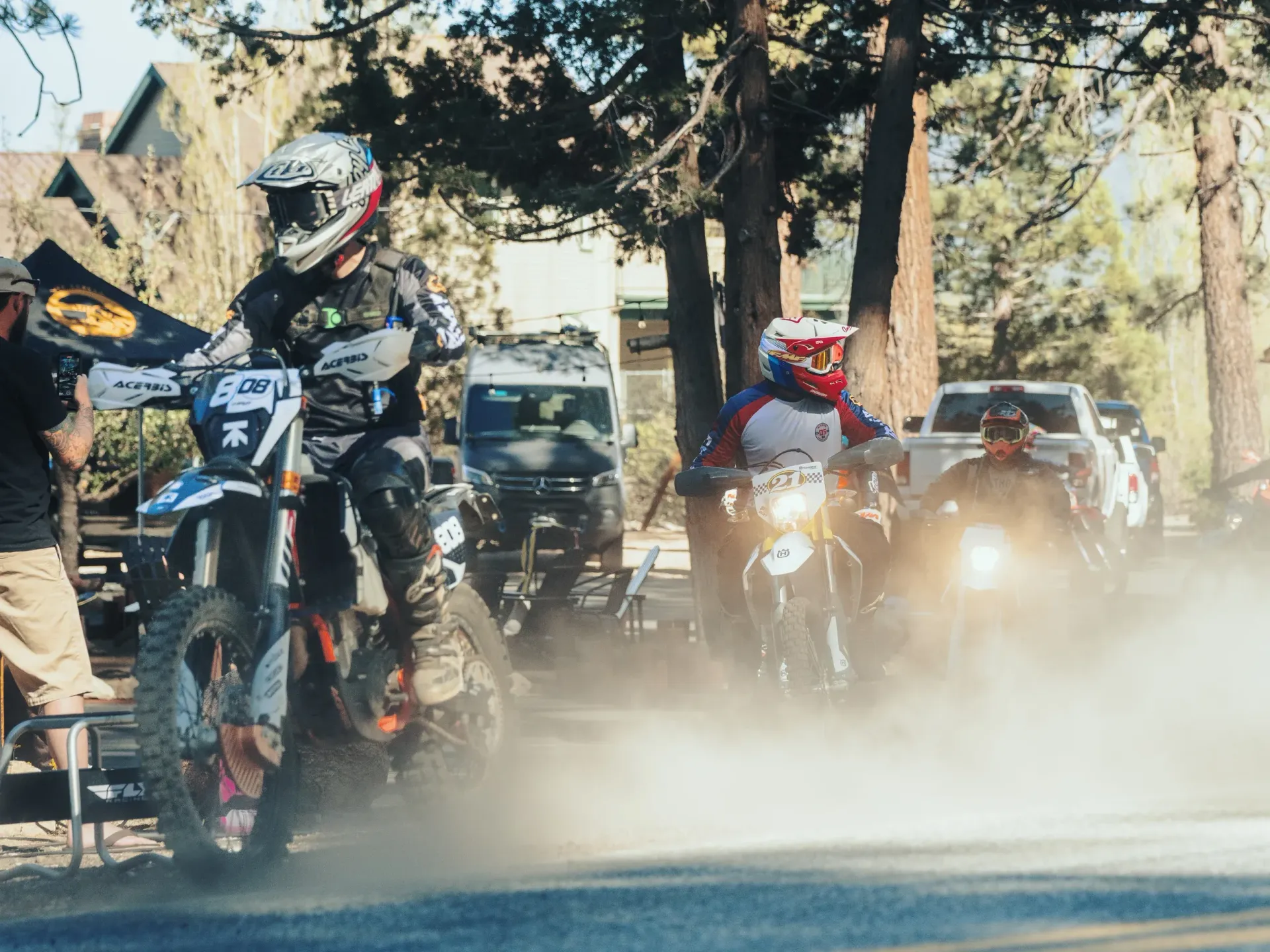 Motorcyclists racing on a dirt road, kicking up dust. Riders wear helmets and protective gear in a wooded area.