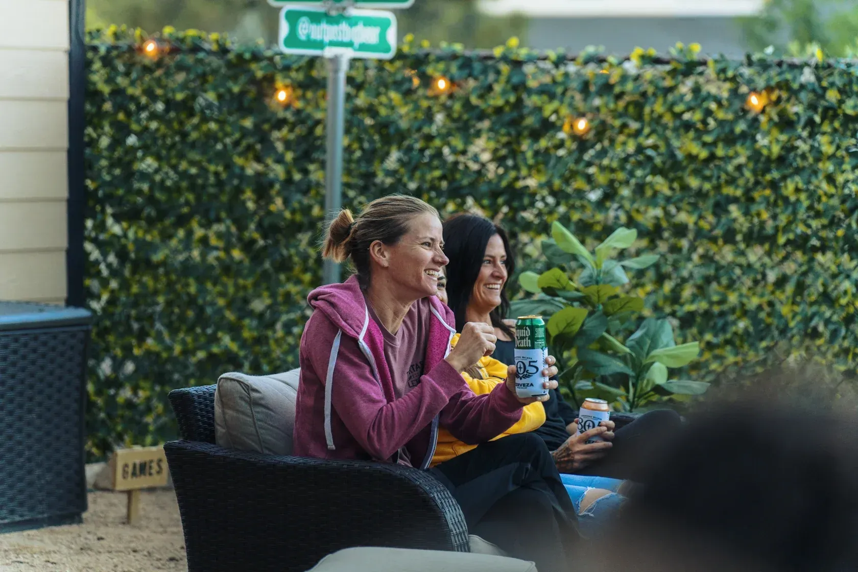 Two women smiling, drinking beer, seated on patio furniture in front of a green wall and string lights.