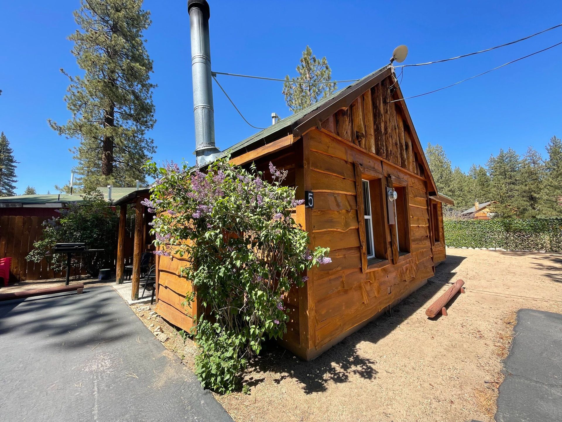 Wooden cabin with a tall chimney on a sunny day. Bush in front, gravel and pavement surround.