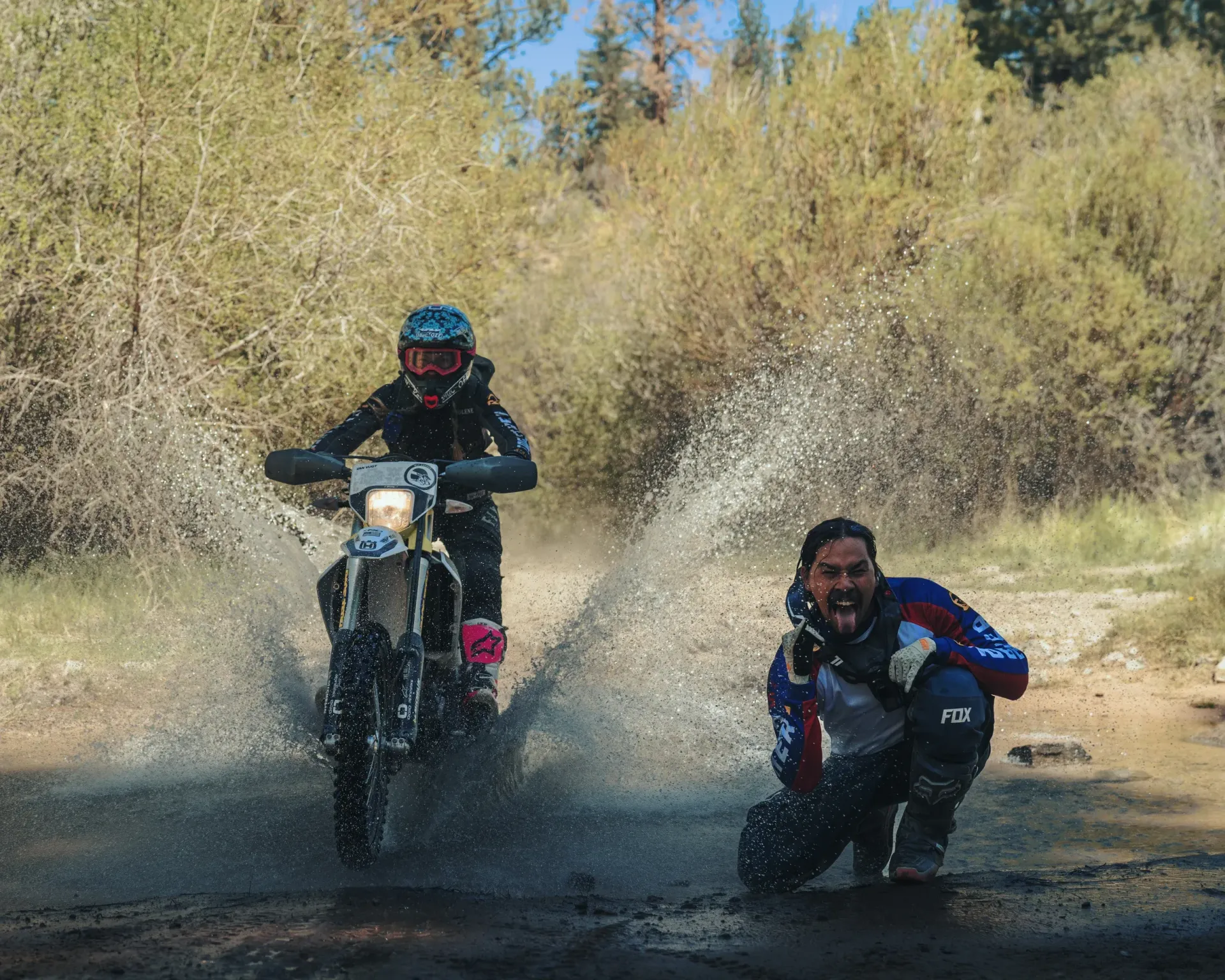 Motorcycle rider splashes through puddle, spraying water. A person crouches to the right, smiling. Outdoors, trees in background.
