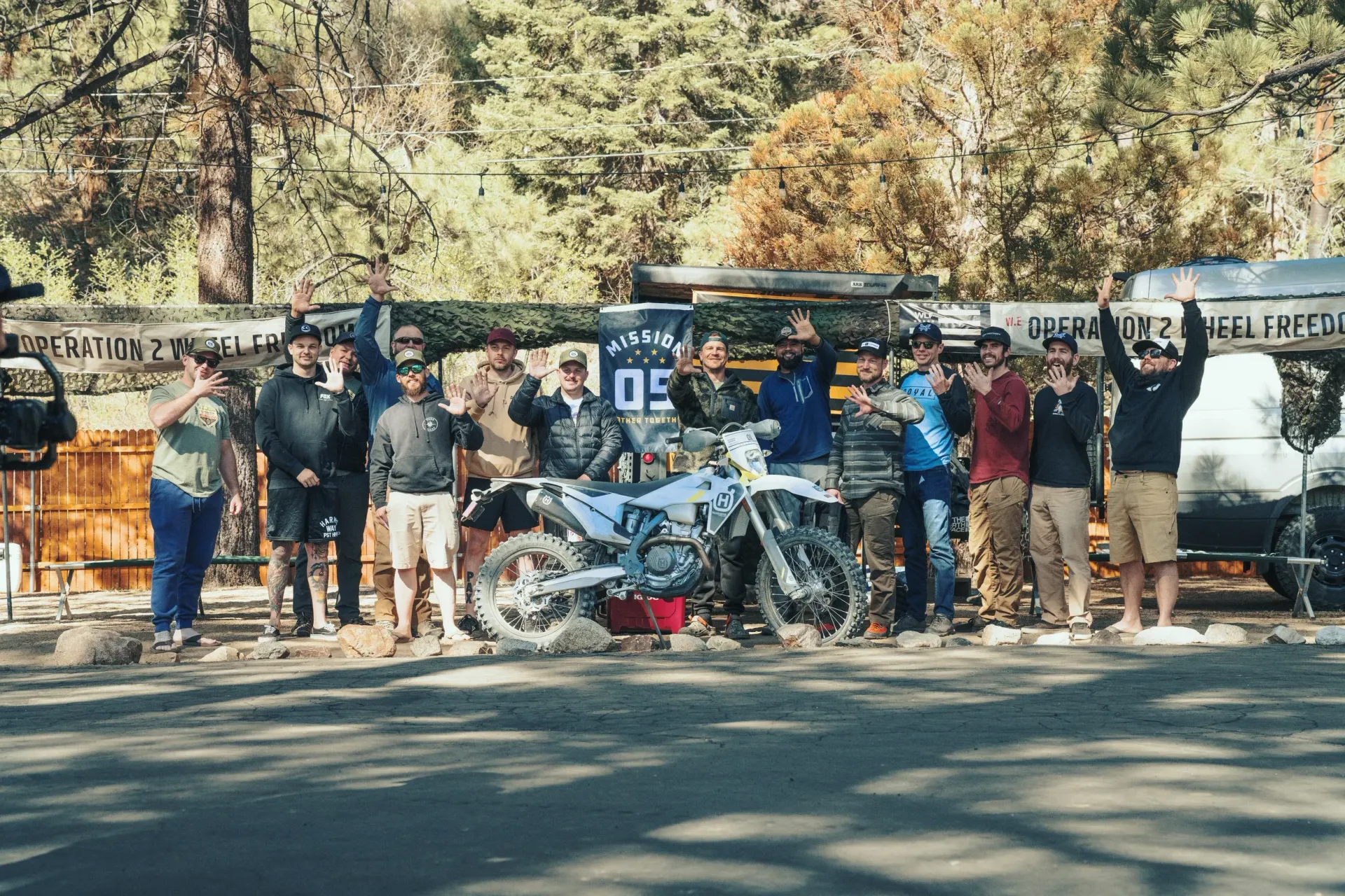 A group of people standing next to a motorcycle, many raising their arms. Trees and a banner are in the background.