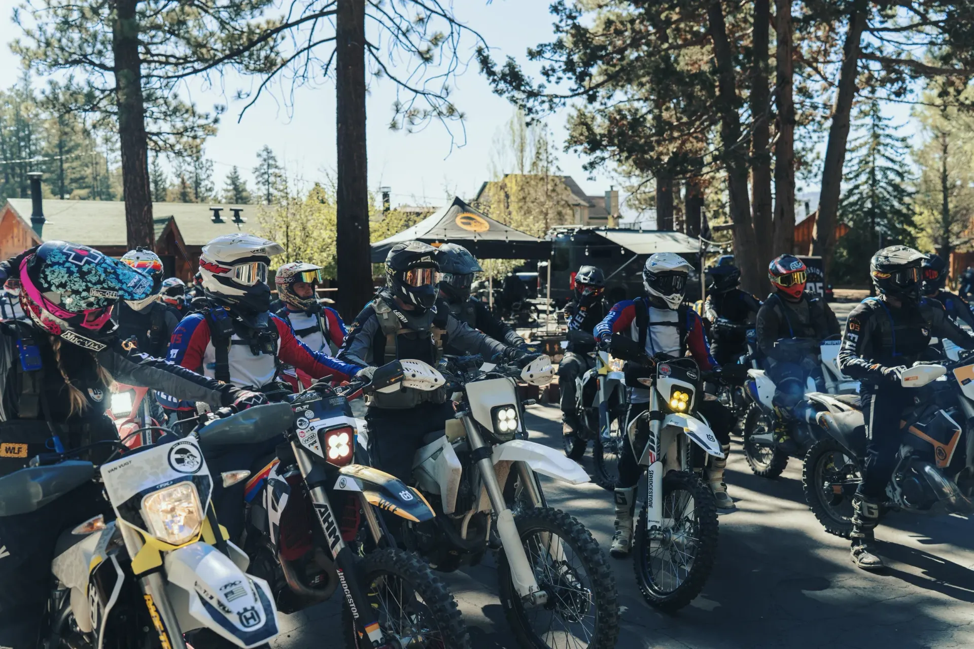 Group of dirt bike riders wearing helmets and gear, lined up on a road, trees and buildings in the background.