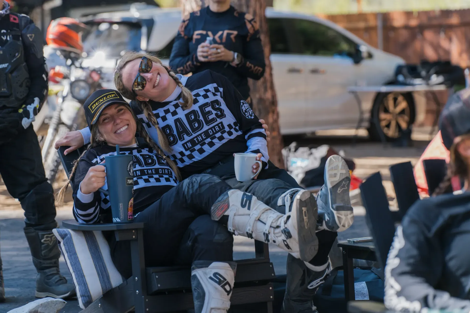 Two women in moto gear relax outdoors, smiling, holding mugs. They sit in chairs, legs crossed, with parked cars behind.