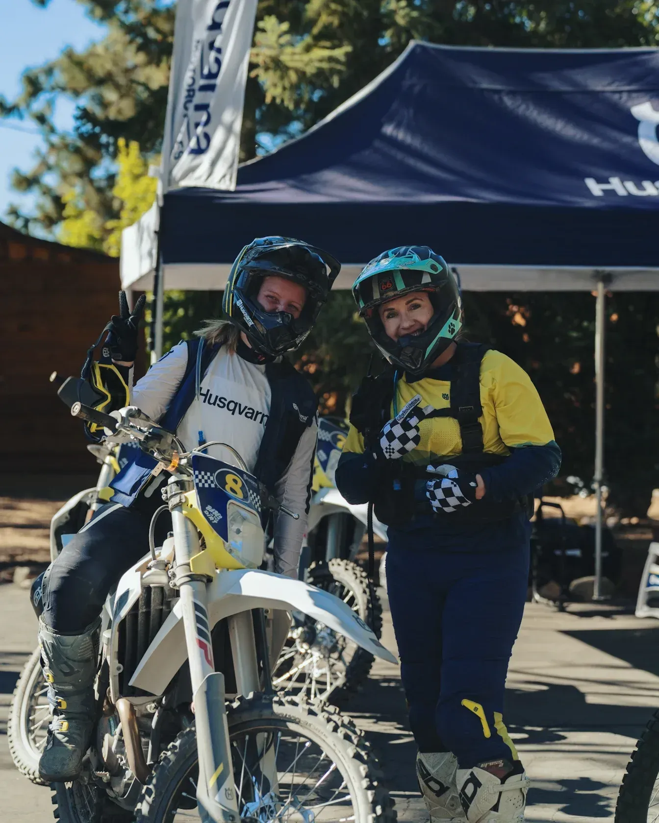 Two people in helmets and riding gear pose by a Husqvarna motorcycle. One sits on the bike, giving a thumbs-up.