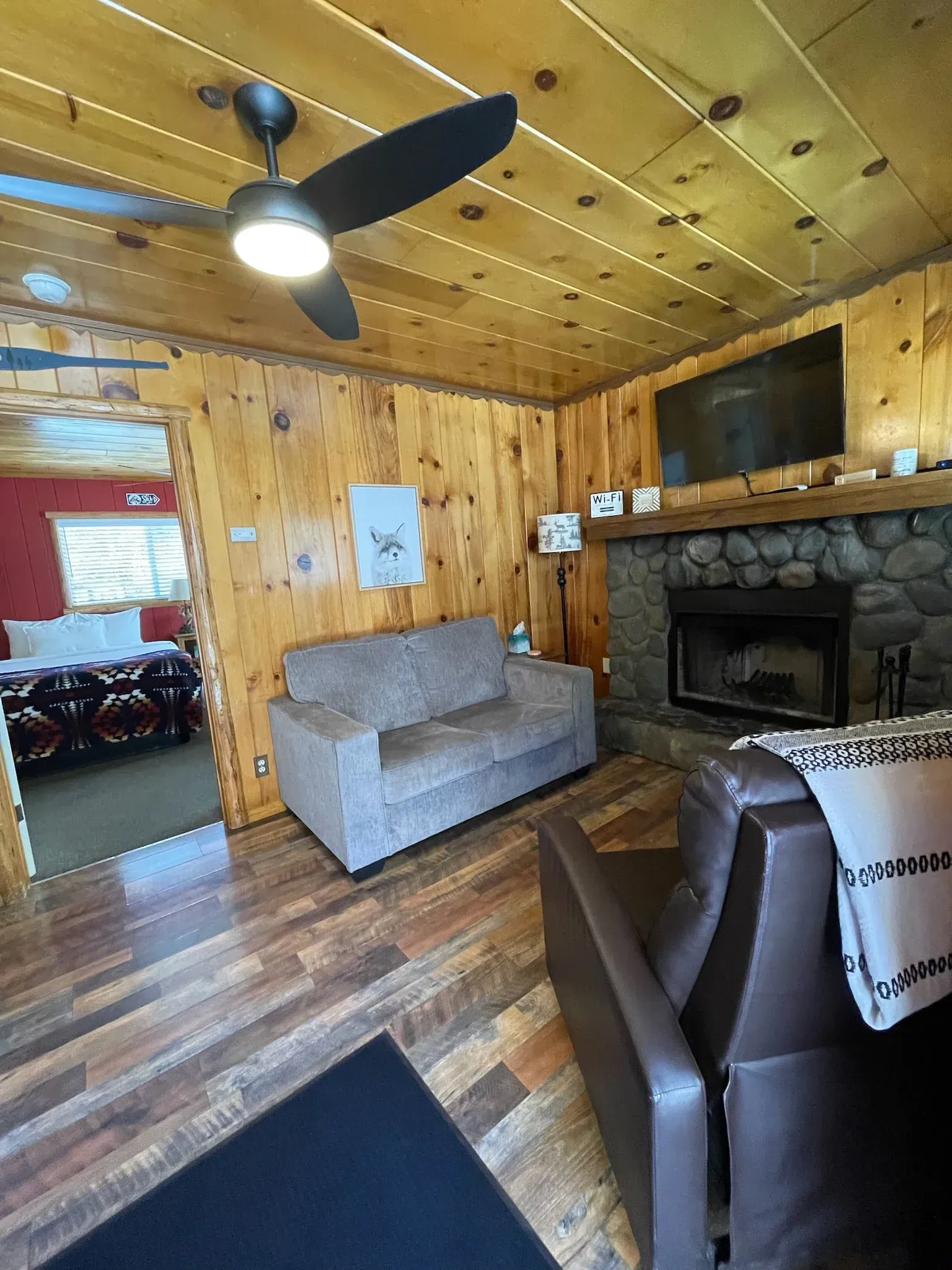 Living room with wood paneling, couch, armchair, fireplace, and bedroom visible through doorway.