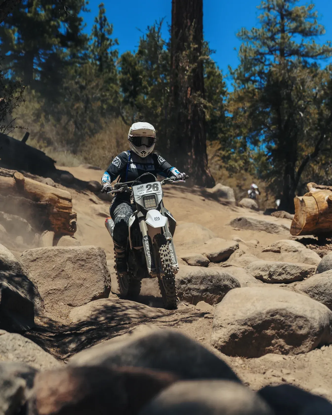 Dirt bike rider navigating a rocky trail, sunny forest setting.