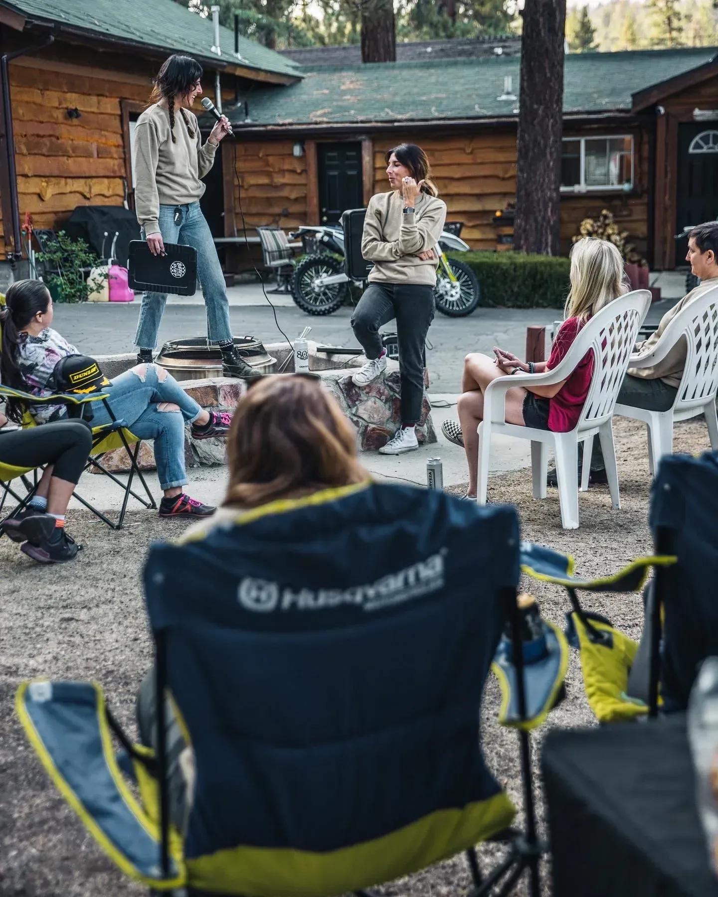 Group of people outdoors in chairs around a fire pit, two people standing and talking.
