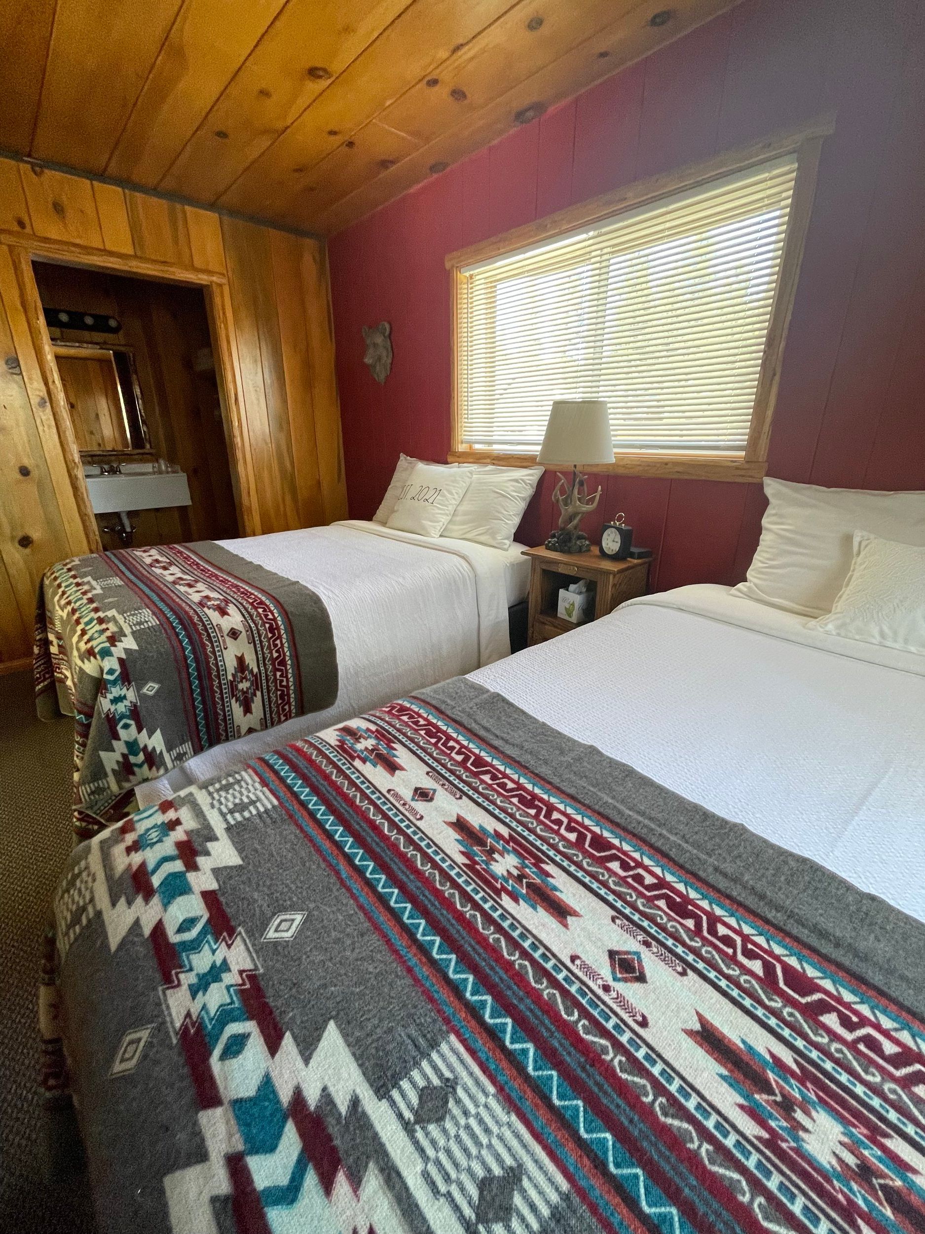 Bedroom with two beds, red walls, patterned blankets, small nightstand, window with blinds, and doorway to bathroom.