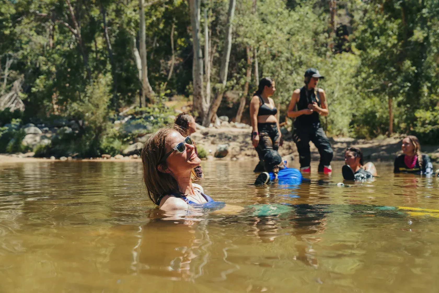 People in a river. One person floats, looking up. Others stand on the bank, one holding a blue bucket. Forest in the background.