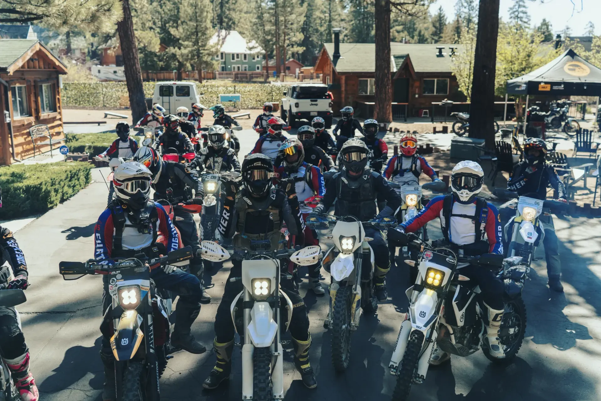 Group of dirt bike riders wearing helmets and gear, lined up on a road, with cabins and trees in the background.