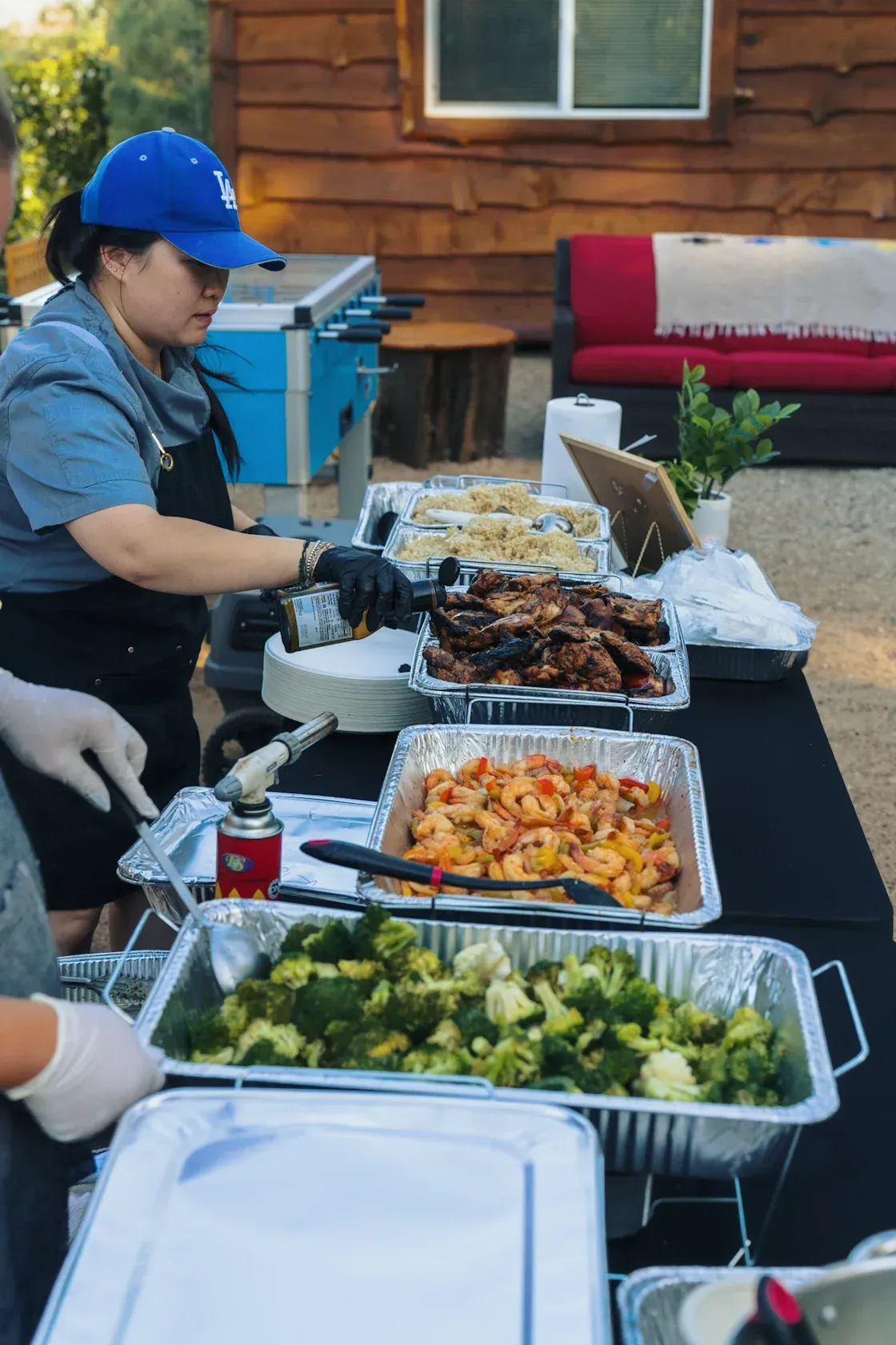 Person serving food from buffet trays at an outdoor gathering. Broccoli, shrimp, and other dishes are visible.