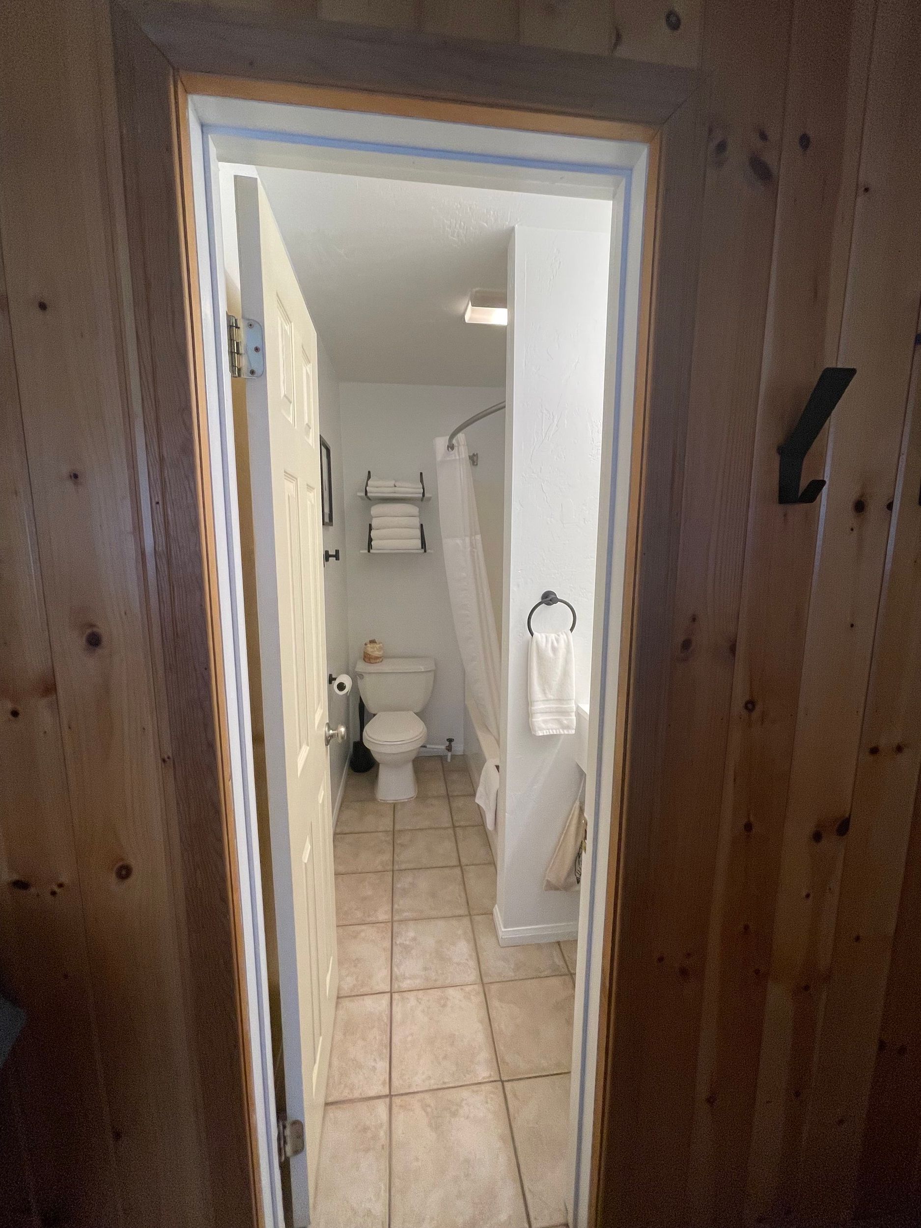 View through a doorway into a bathroom with a toilet, shower, and towels. Light wood paneling surrounds the door frame.