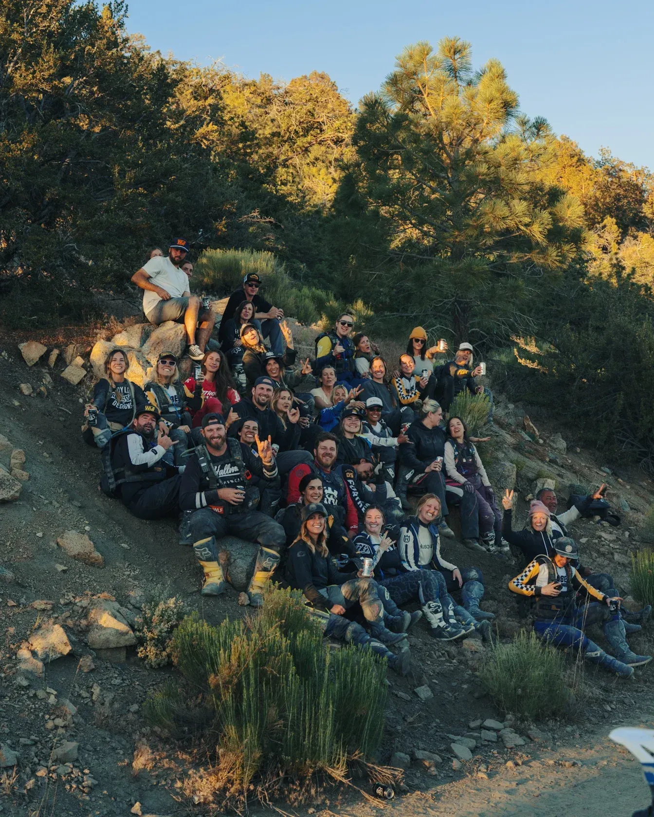 Group of people, many in motorcycle gear, posing together on a hillside in the sunlight.