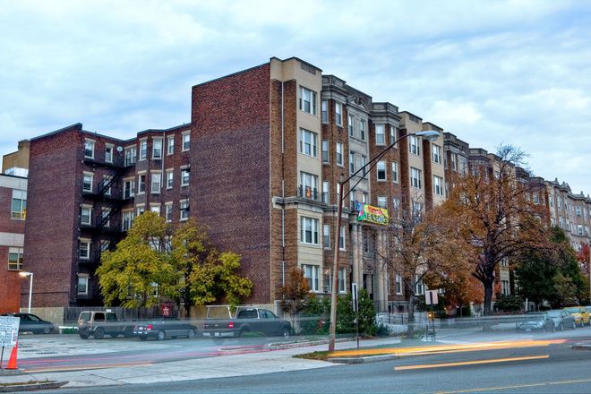 Multi-story brick apartment building on a cloudy day; street level with trees and parked vehicles.