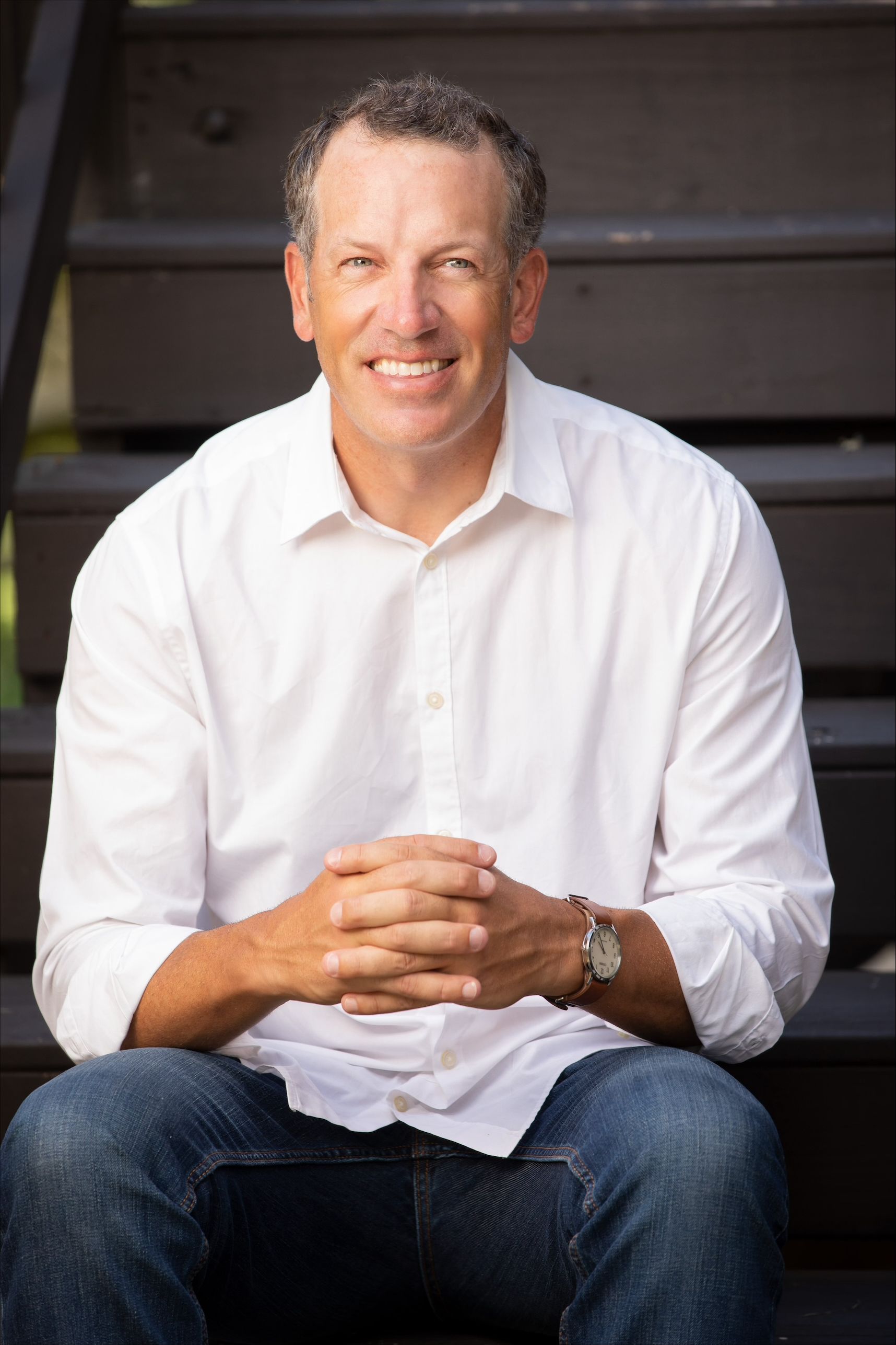 A man in a white shirt is sitting on a set of stairs with his hands folded and smiling.