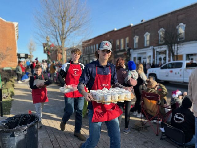 Three young men in red aprons carry drinks to people on a sunny street.