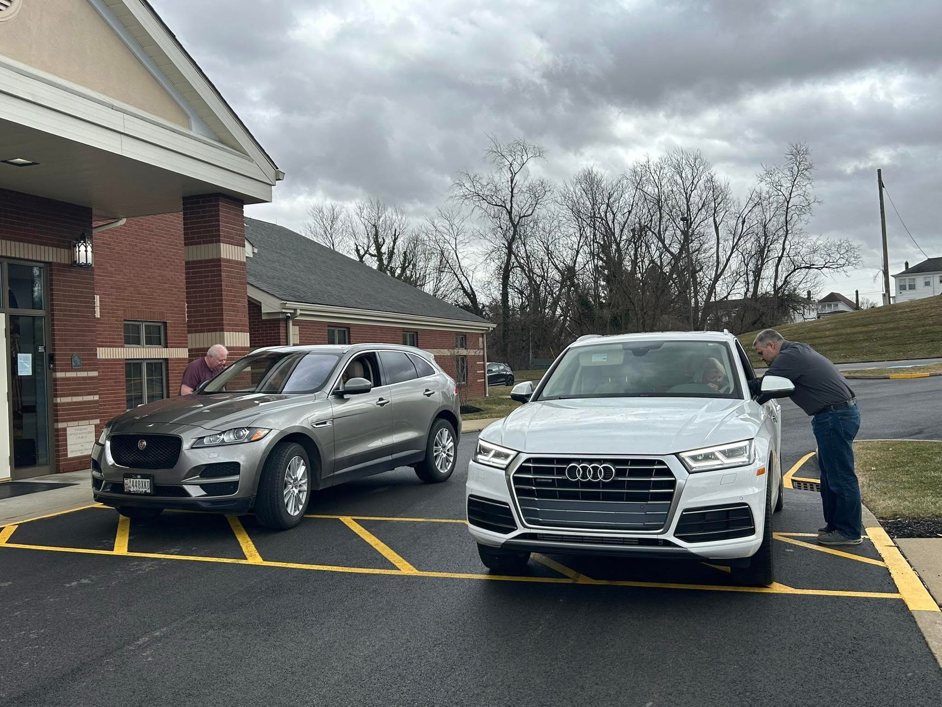 A gray Jaguar and white Audi parked in front of a brick building. A person is leaning towards the Audi.