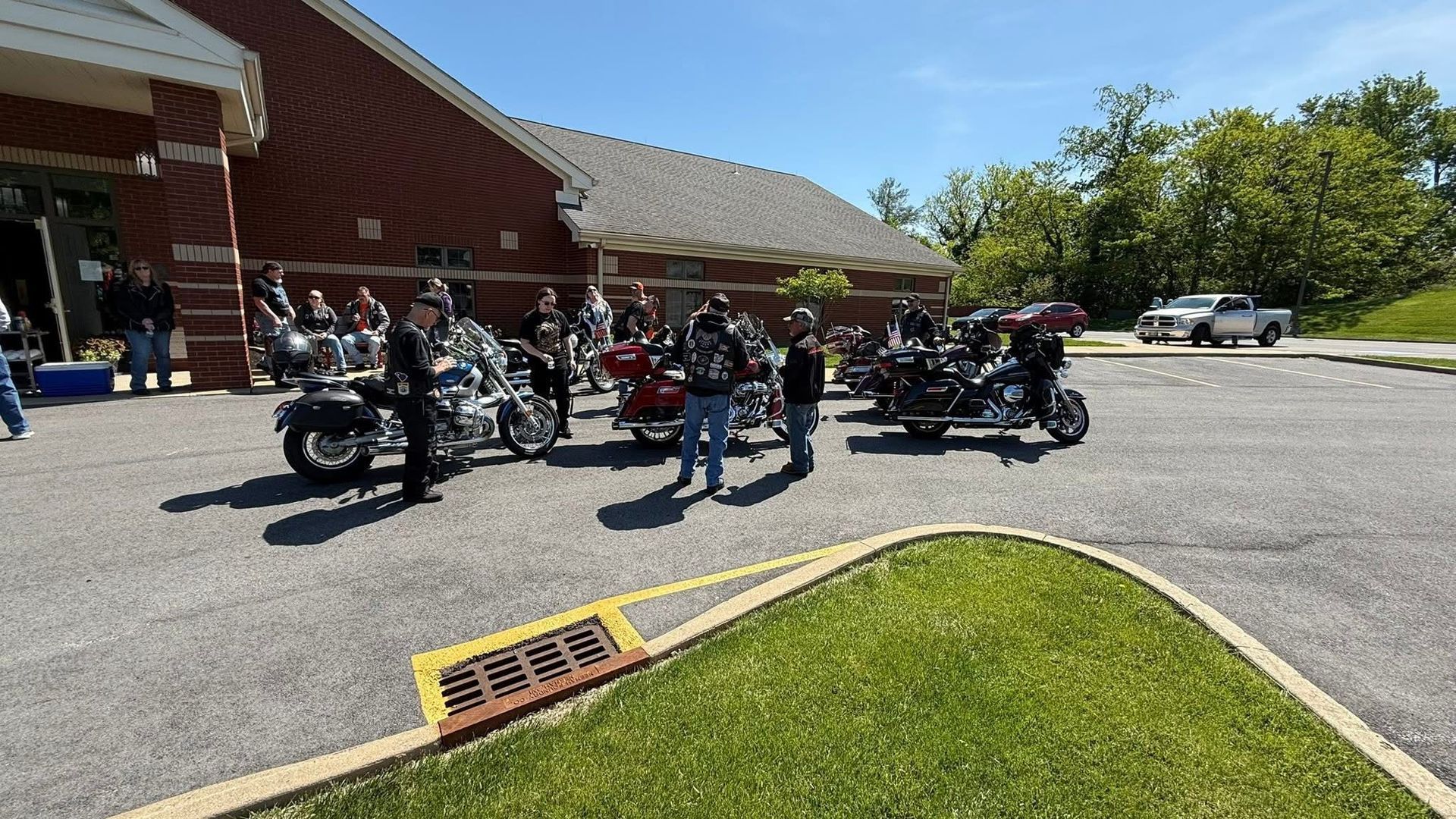 Motorcycles parked outside a brick building; people standing around, sunny day.