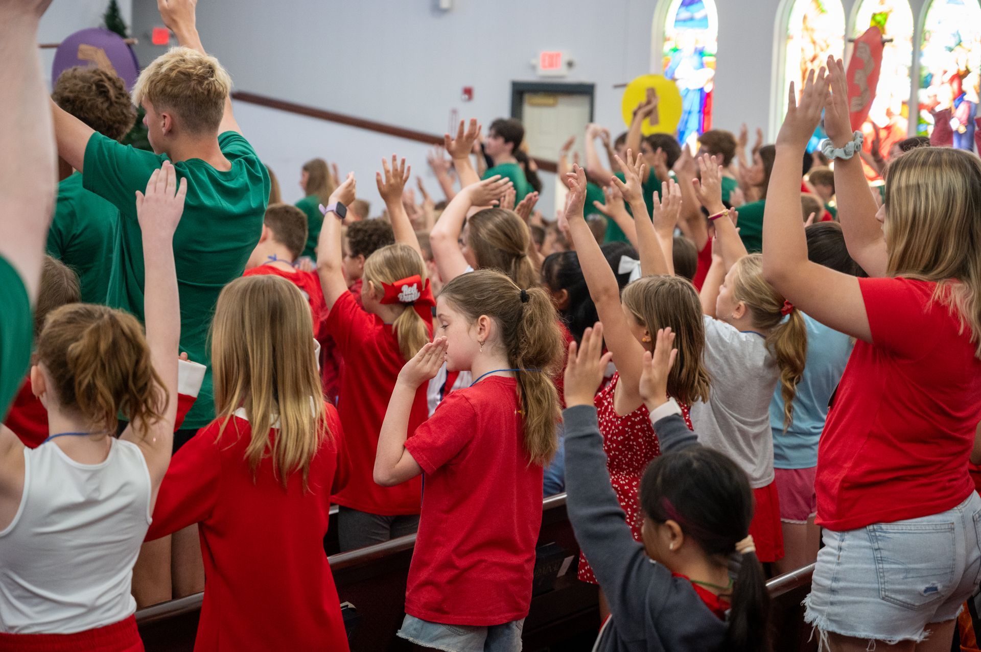 Children in red and green shirts raise their hands in a church, possibly singing or worshiping.