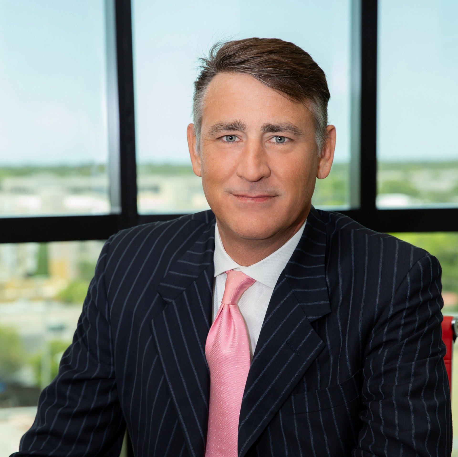 A man in a suit and pink tie is sitting in front of a window.