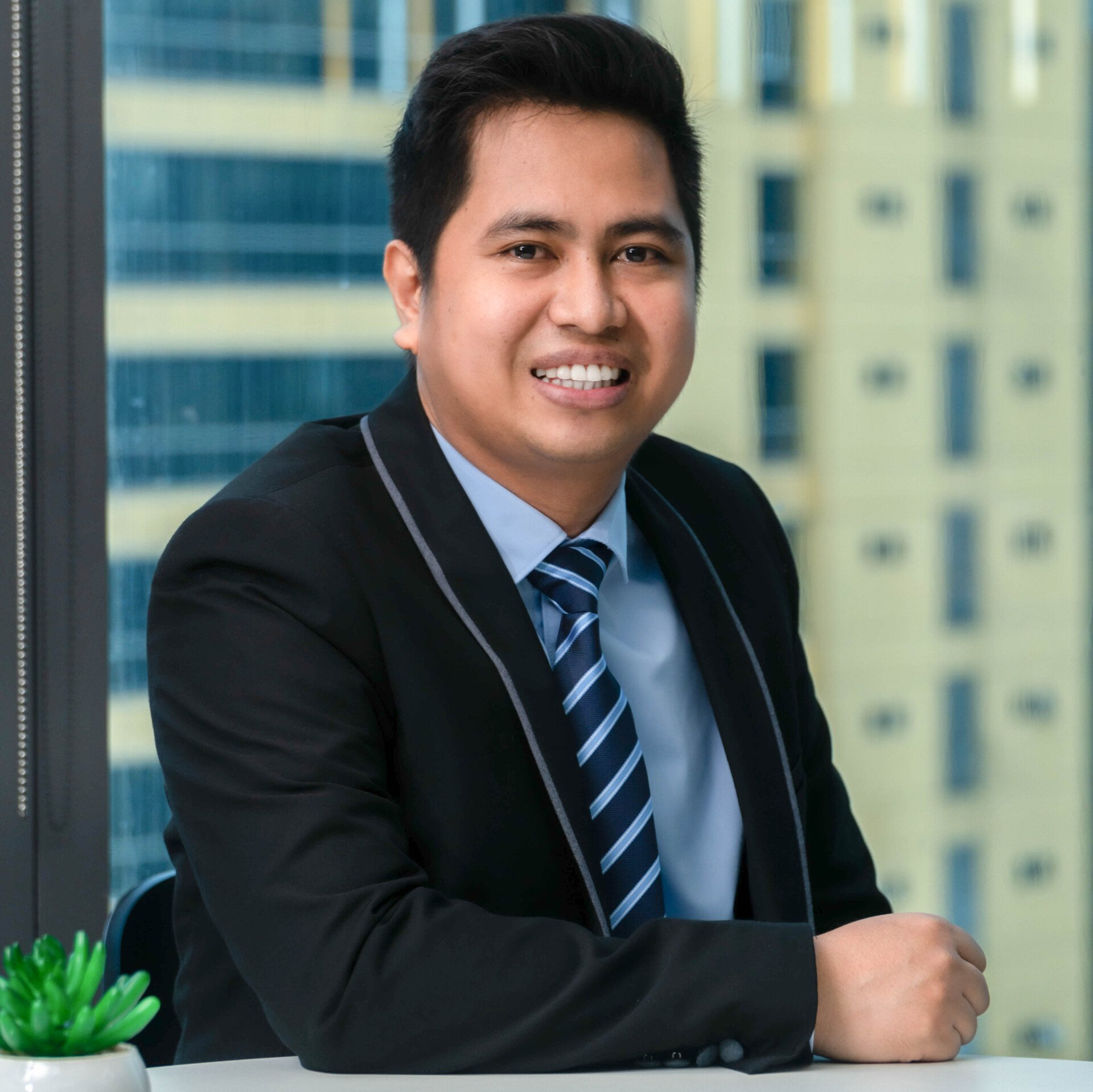 A man in a suit and pink tie is sitting in front of a window.