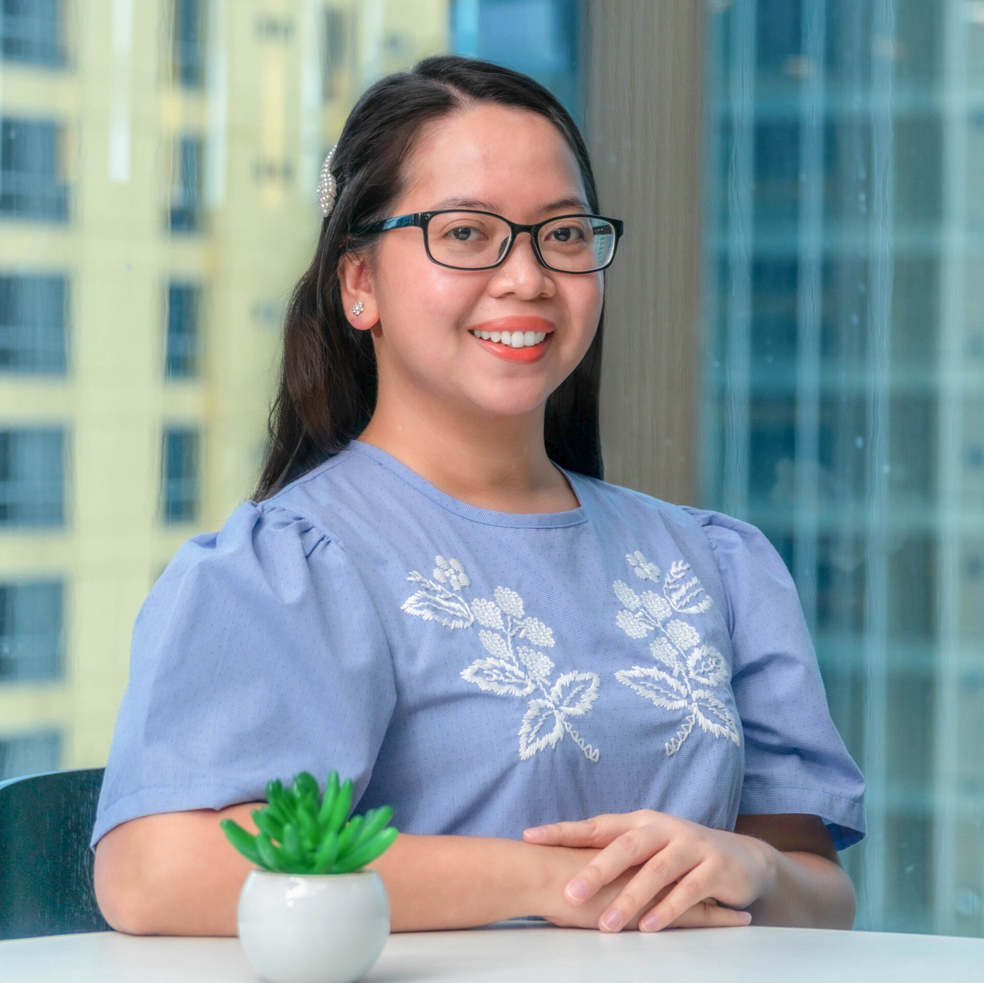 A woman wearing glasses is sitting at a table with a potted plant.