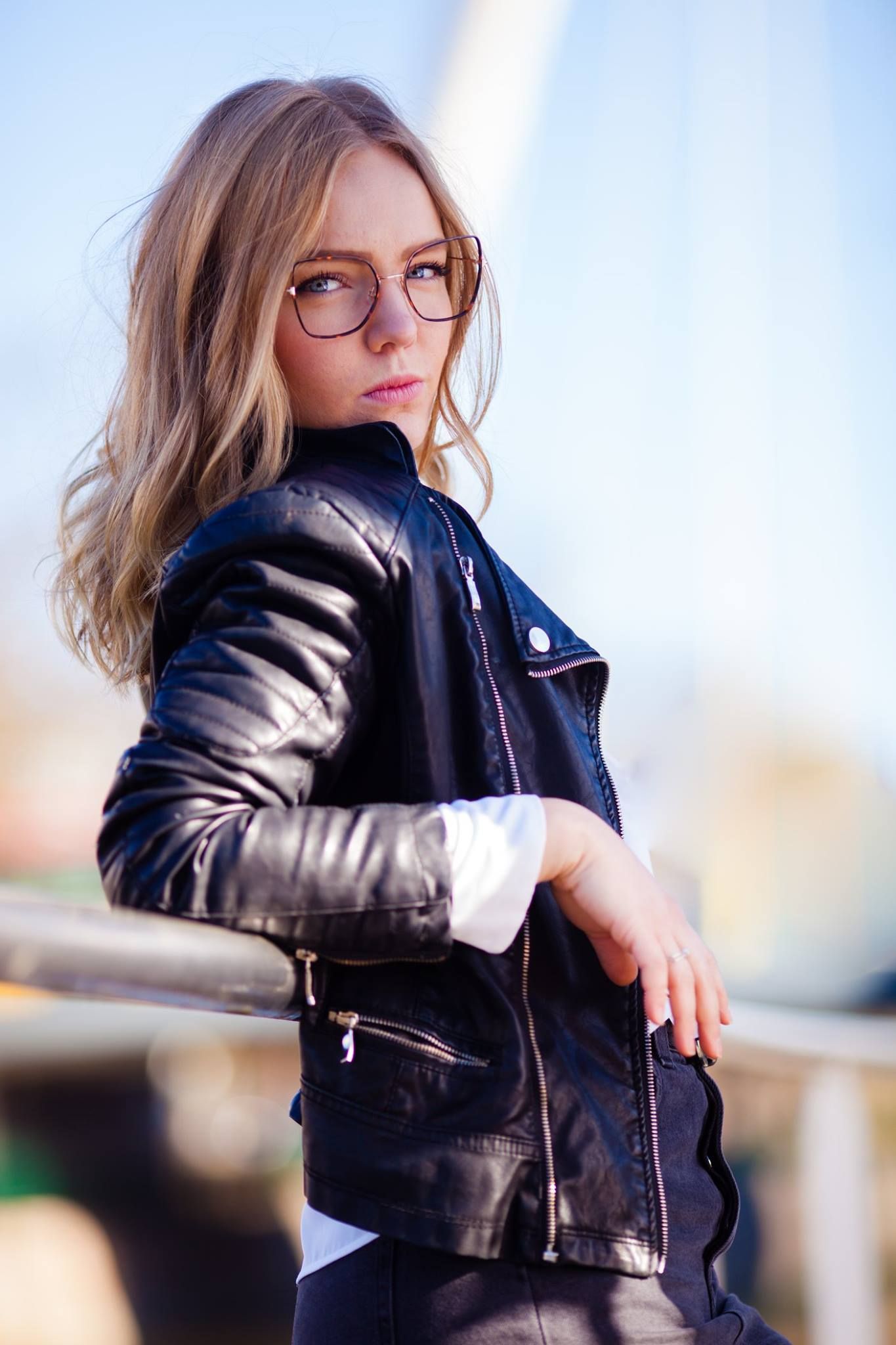 Une jeune femme portant des lunettes et une veste en cuir est accoudée à une balustrade.