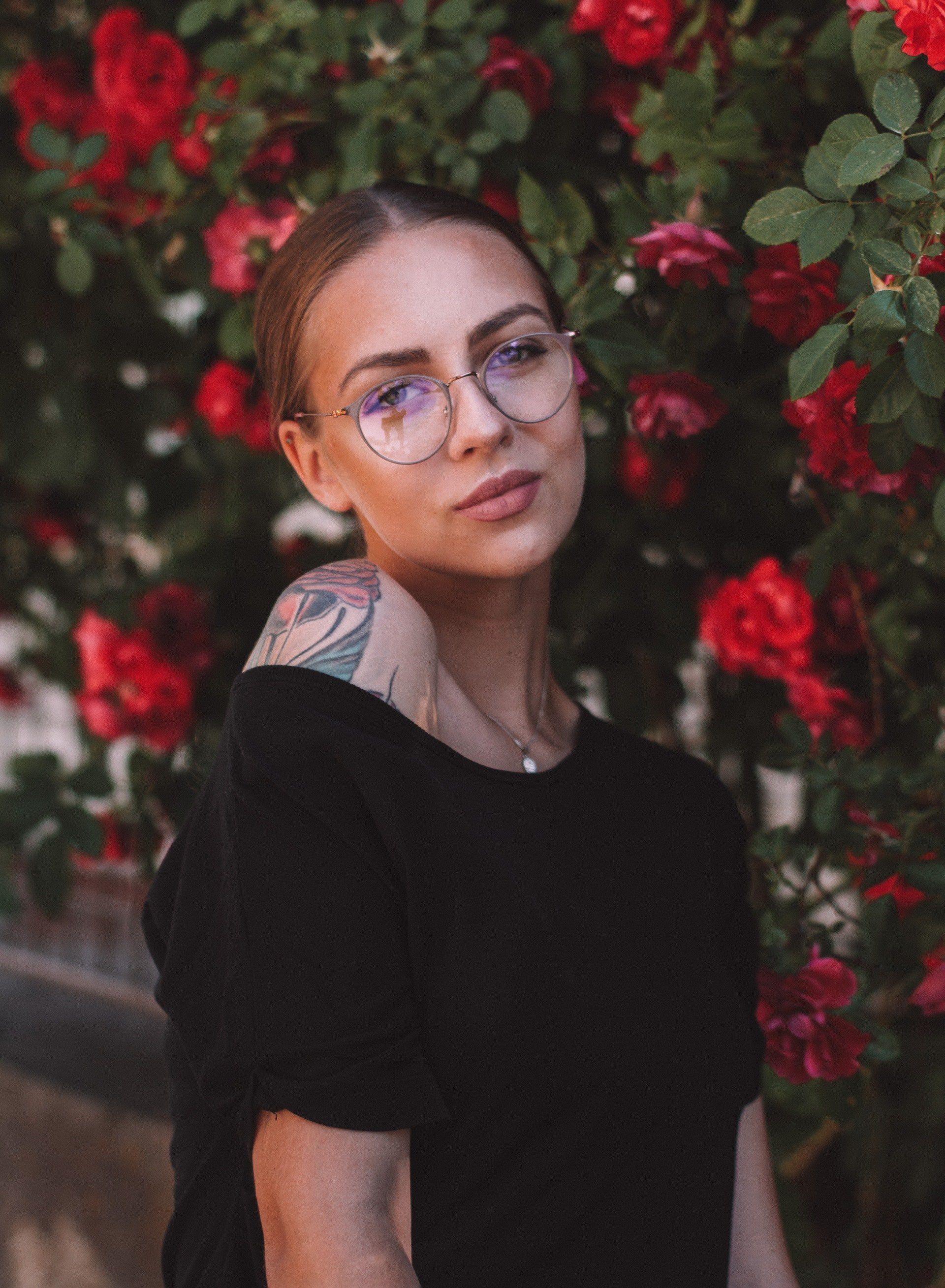 Une femme portant des lunettes et une chemise noire se tient devant un buisson de roses rouges.
