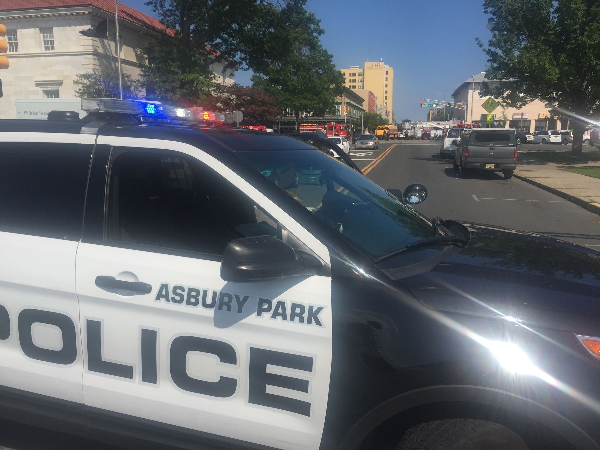 Asbury Park Police car with flashing lights on a street, likely a crime scene, in daylight.