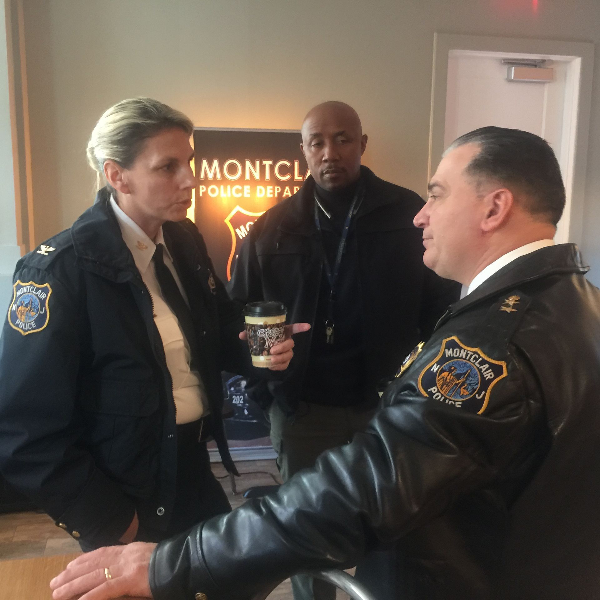 Three uniformed police officers in an office, discussing, one holding a coffee cup. 