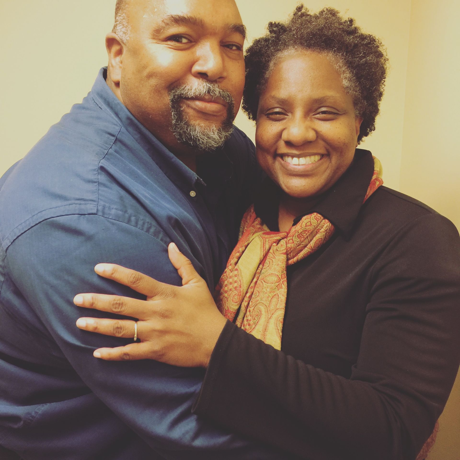 Smiling couple embraces; man in blue shirt, woman in black shirt with scarf, both indoors.