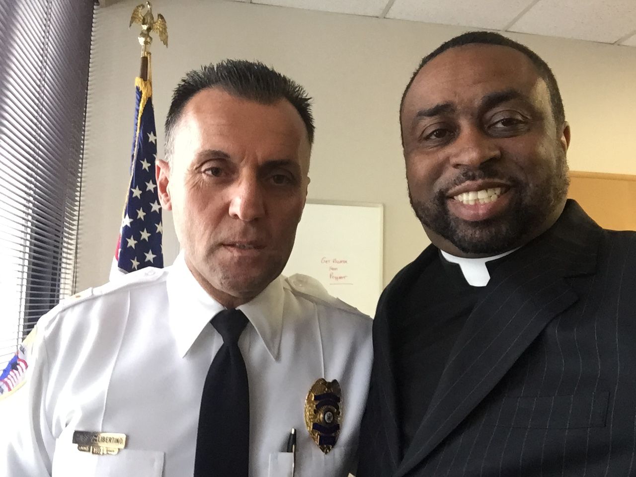 Two men posing. One in uniform, the other in a suit. American flag visible in the background.