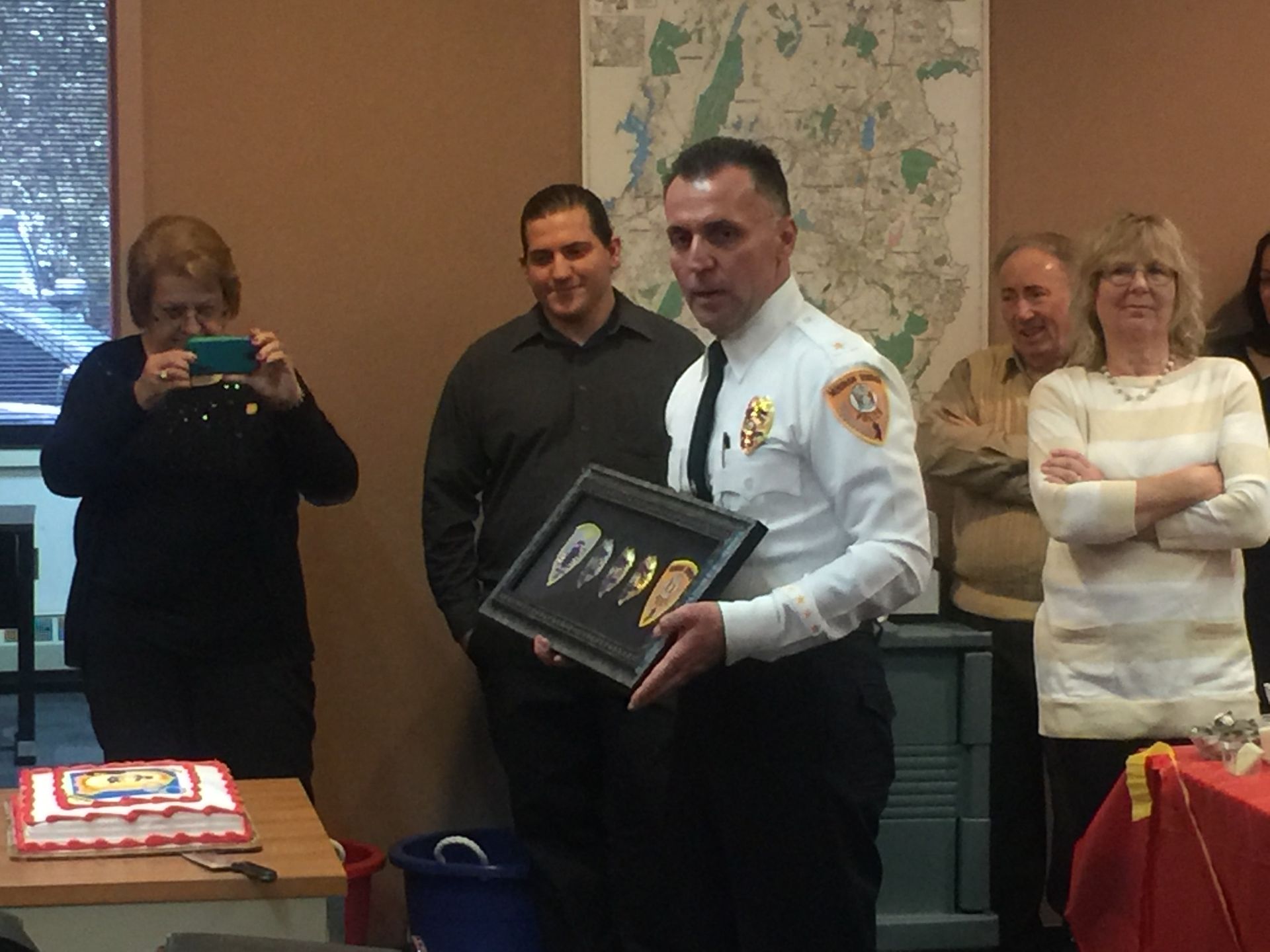 Firefighter holding framed badges; people watch. Cake on a table. Indoor setting.