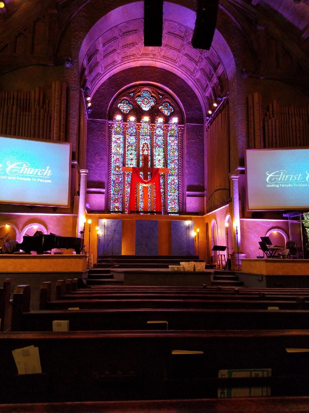Interior of a church with purple lighting, a stained-glass window, and a large red cross. Rows of pews face the stage.