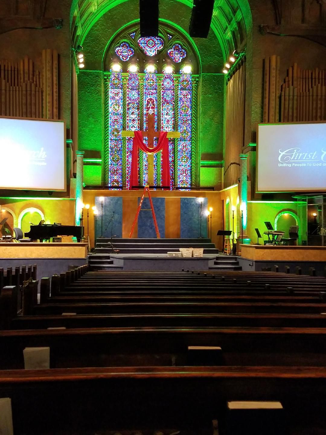 Interior of a church sanctuary with a large stained-glass window and a red cross. Green lighting illuminates the space.
