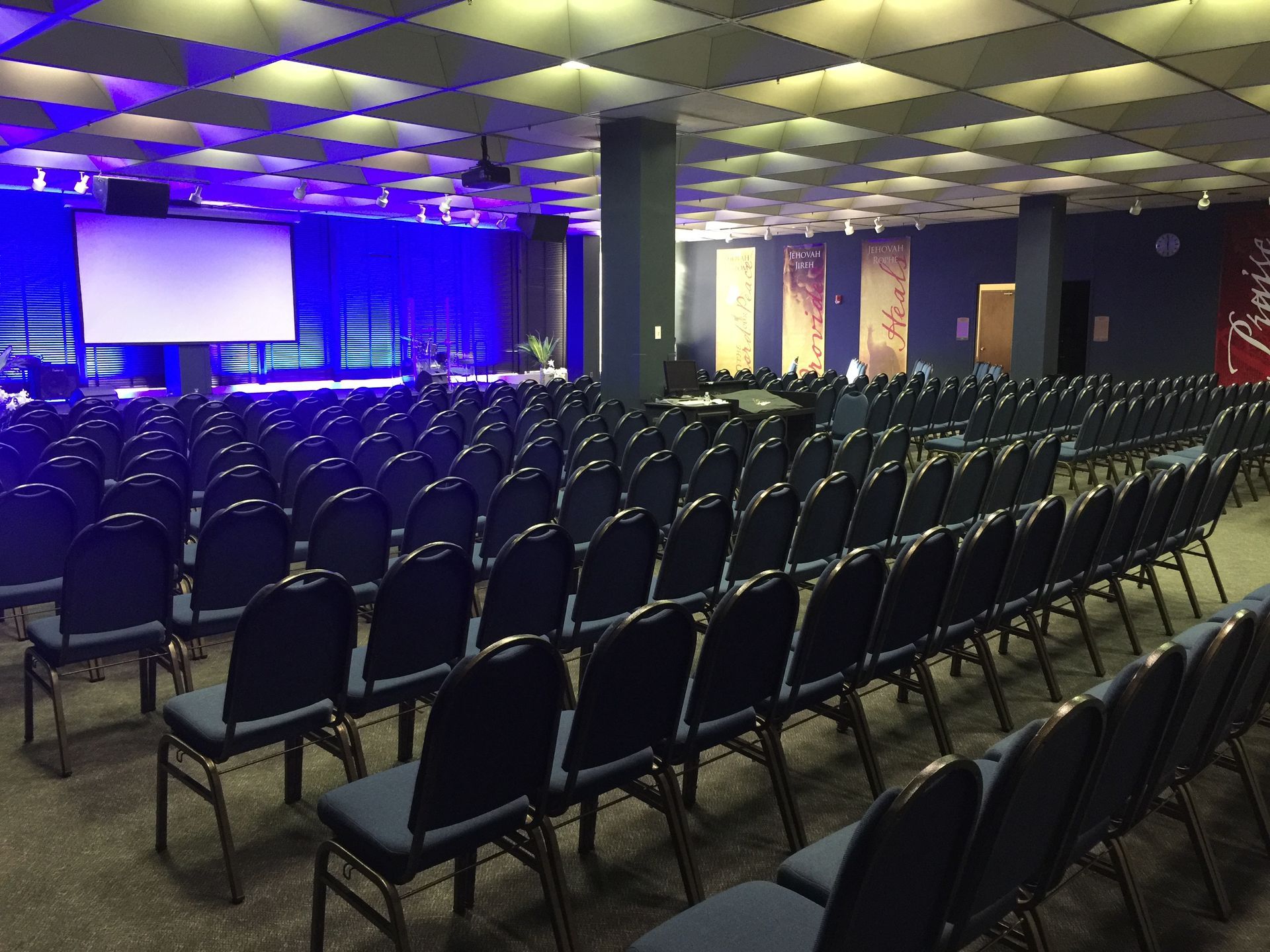 Rows of blue chairs face a stage, lit with blue lights, in a large room.