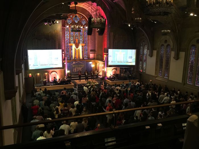 A large church service in progress. People seated, screens up front, stage with performers, colorful lights.