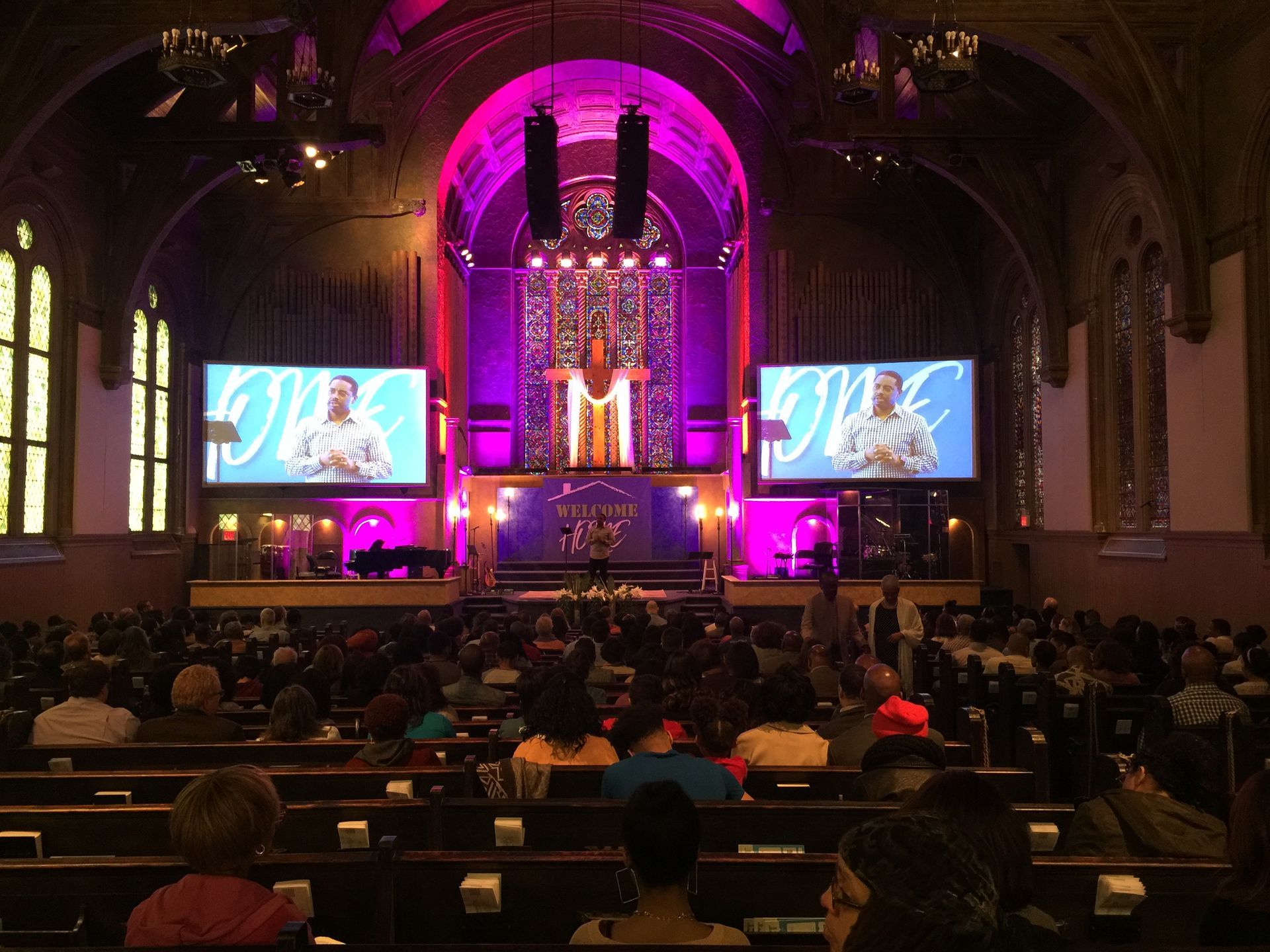 Church interior with audience, stage lit in purple, two screens displaying a person speaking.