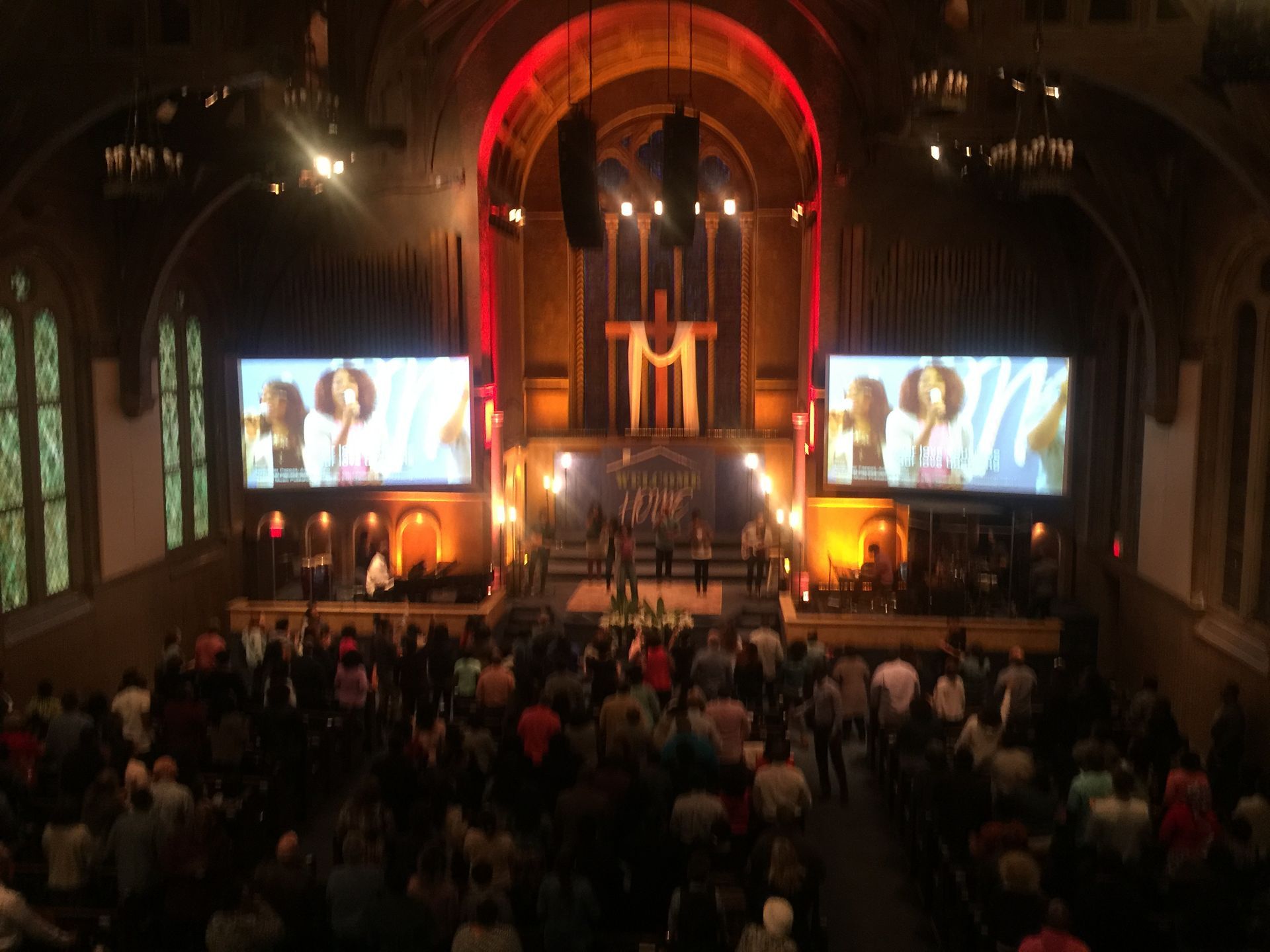 Large church service with screens displaying a singer, stage with musicians, and a crowd of people.