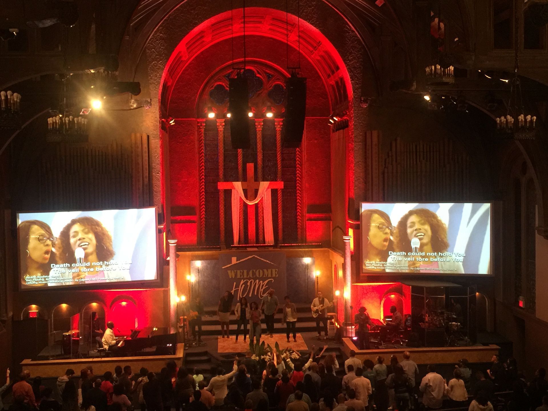 Church service with singers on stage, screens showing the singers, red lighting, and an audience raising hands.
