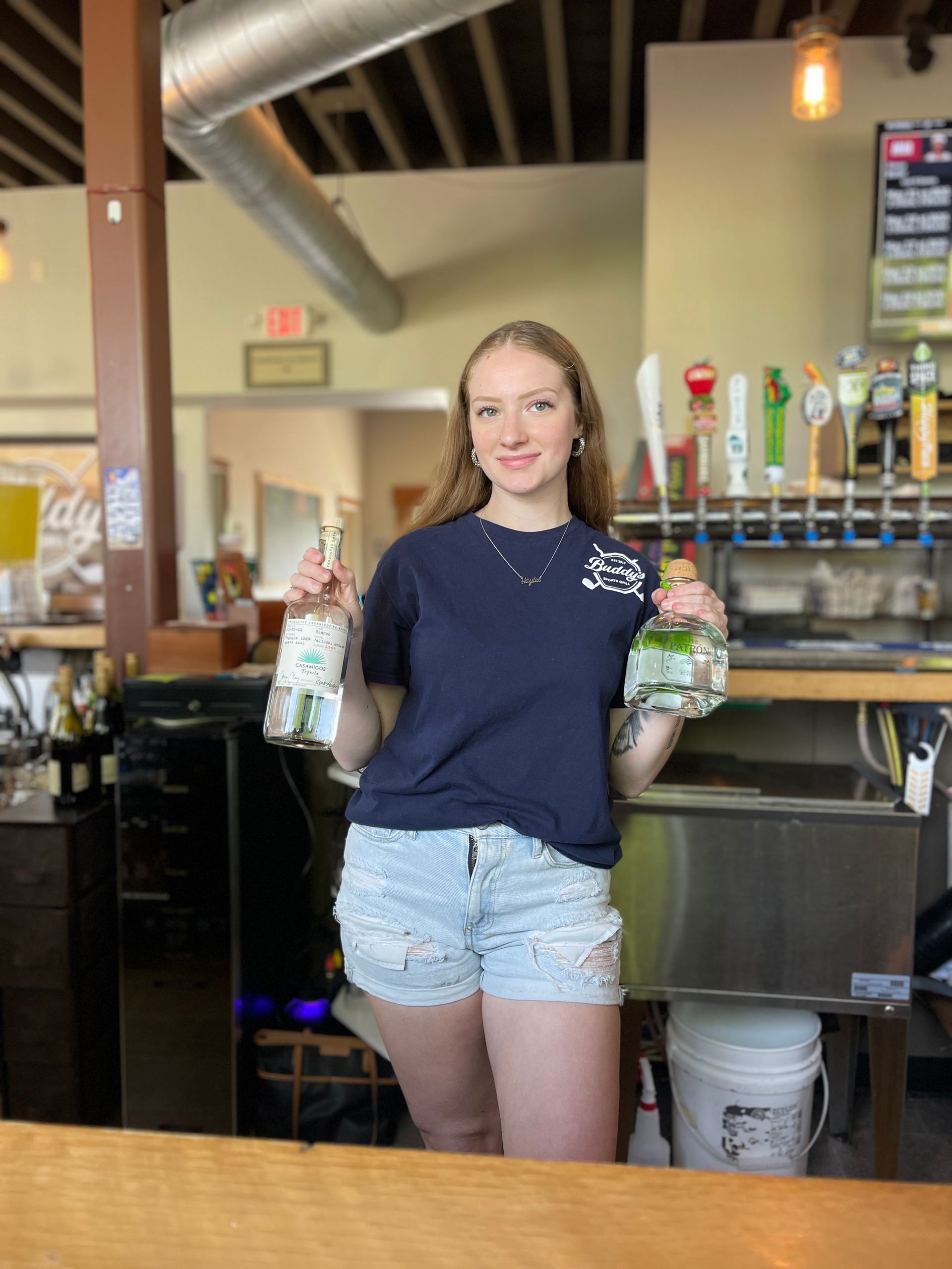 A woman is standing at a bar holding two bottles of beer.